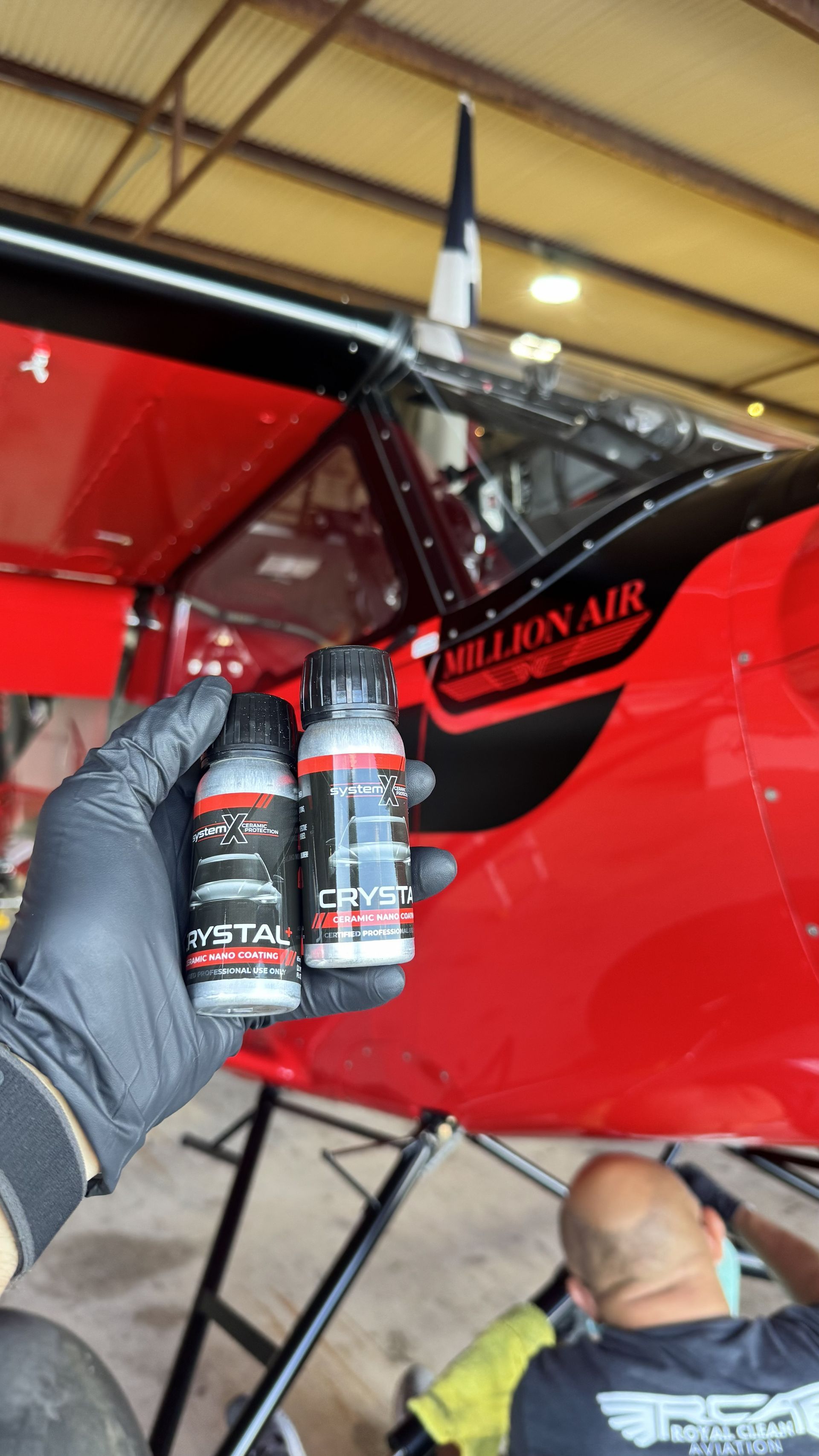 Gloved hand holding two black spray cans beside a red airplane in a hangar