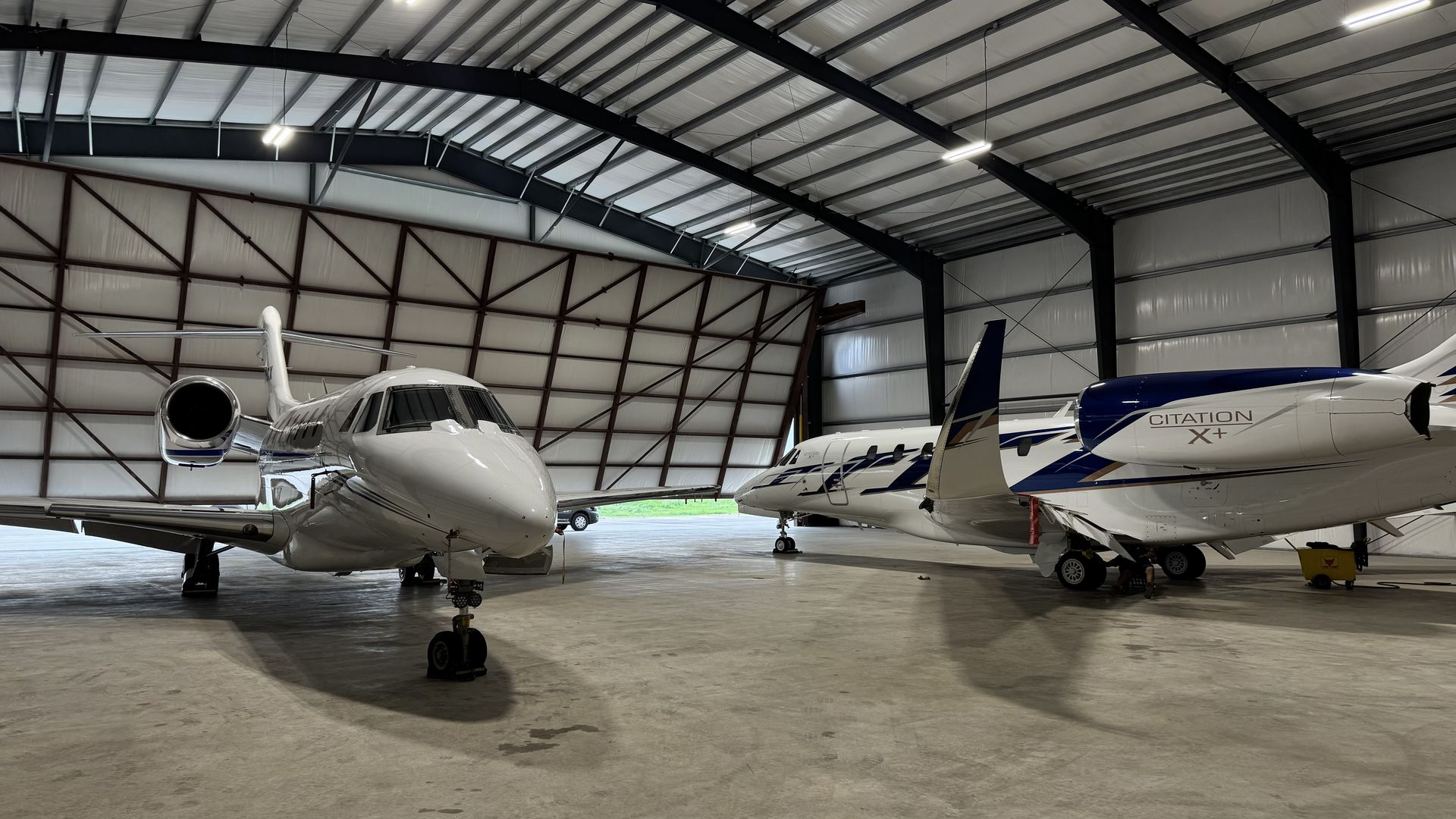 Two white private jets parked inside a large aircraft hangar with a metal roof.