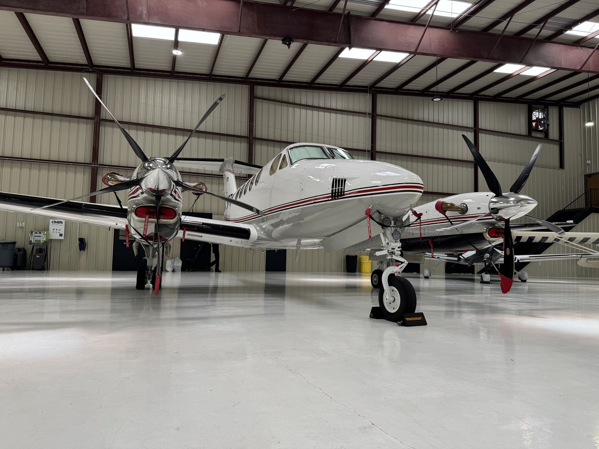 Twin-engine propeller airplane in a hangar with a polished white floor and another aircraft behind it