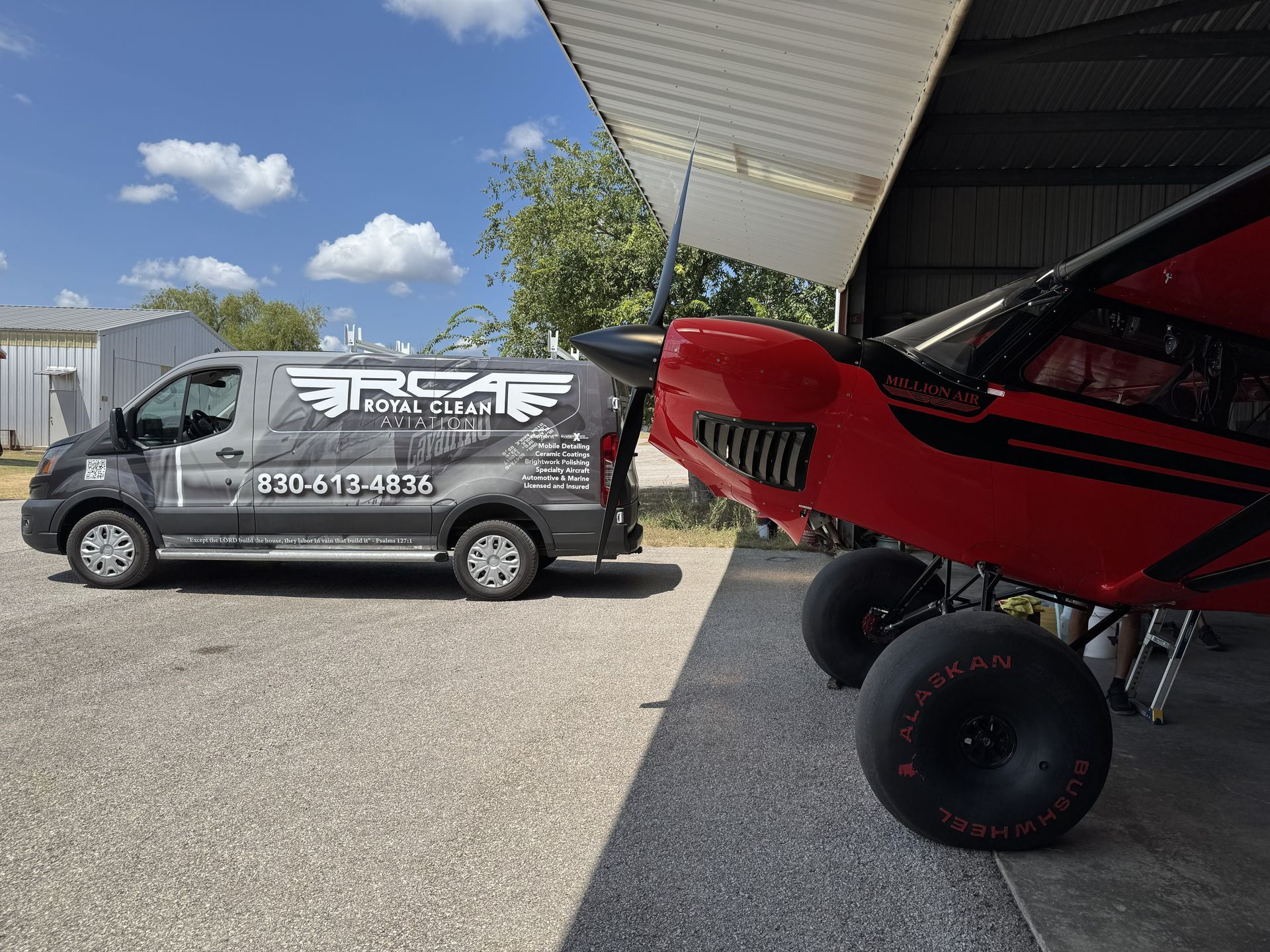 Red off-road airplane parked beside a gray service van under a hangar roof on a sunny airfield