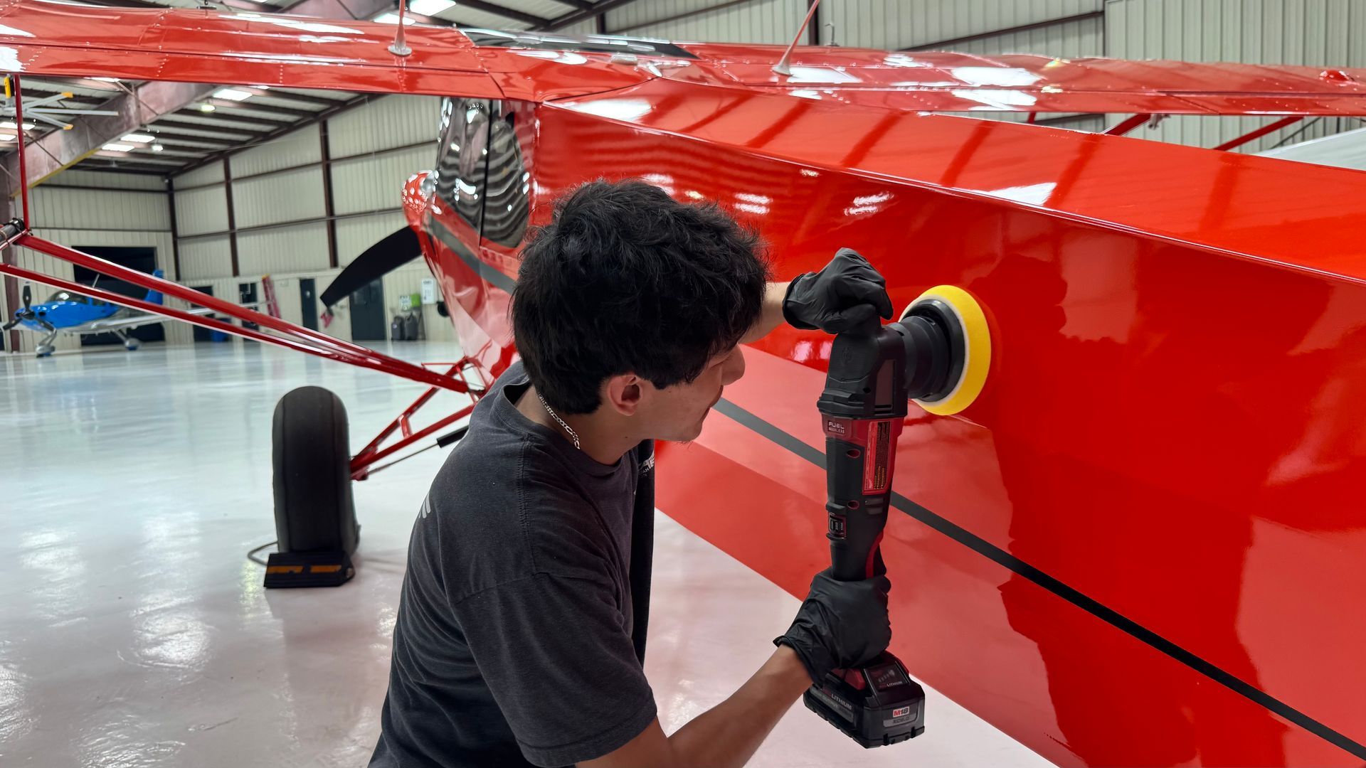 Person polishing a red airplane wing in a hangar with a power buffer
