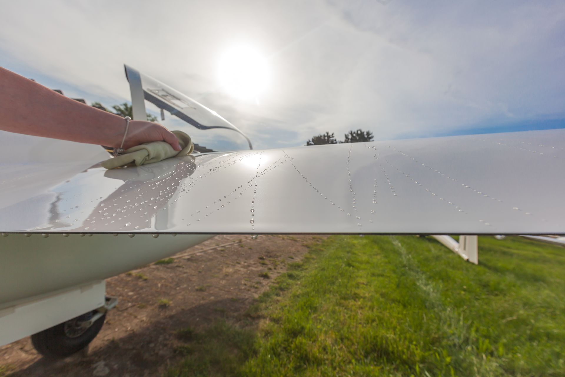Close-up of a white airplane wing with a hand holding the edge, grassy field and sky in the background.