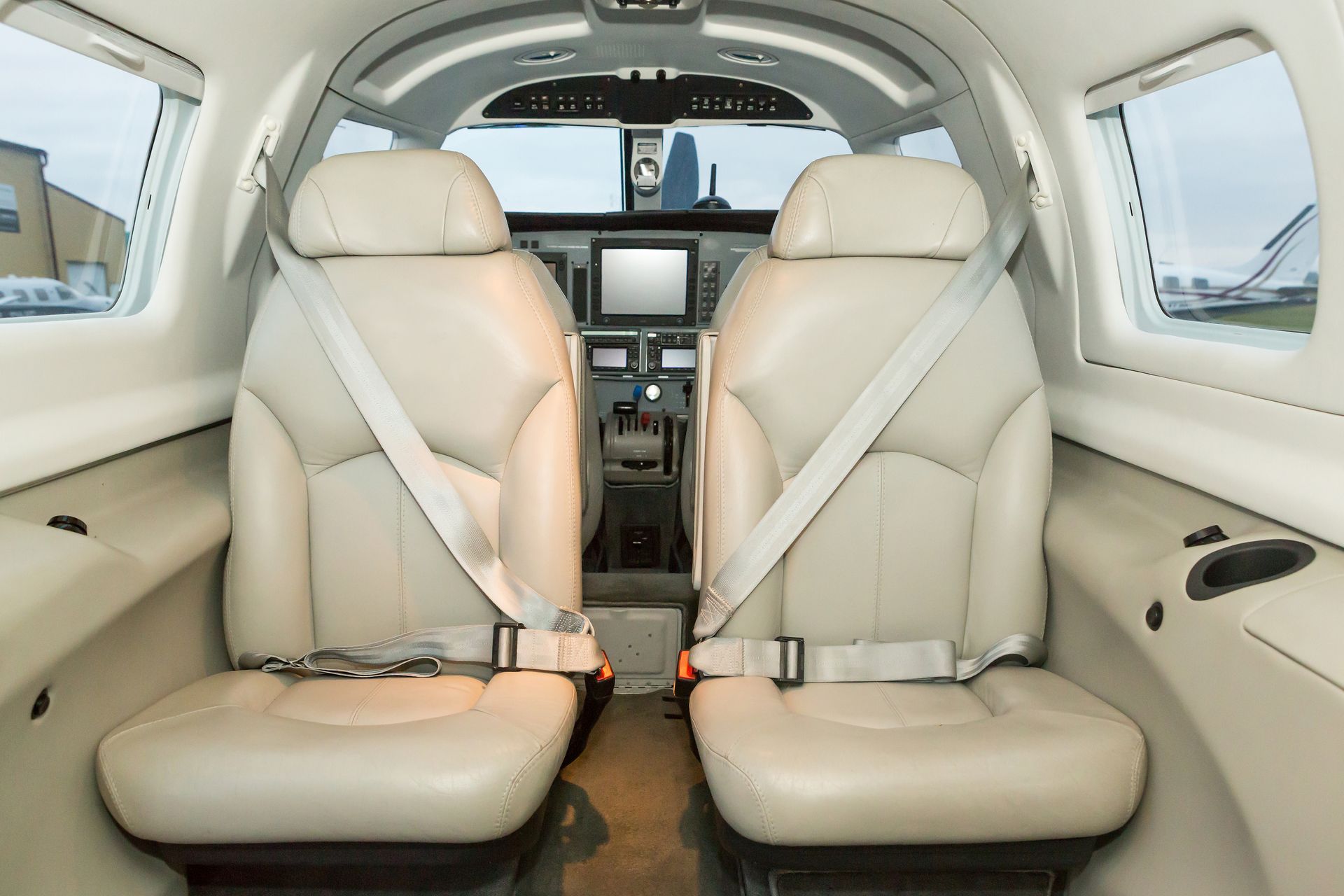Interior of a small aircraft cabin with two beige leather seats, seatbelts, and cockpit visible ahead
