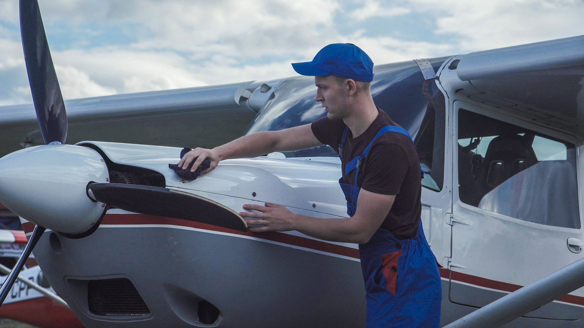 Man in blue overalls and cap working on a small white airplane’s engine on the tarmac
