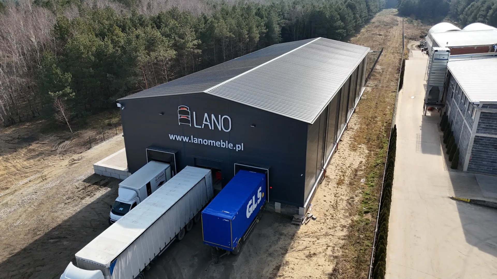 Widok z lotu ptaka na szary magazyn oznaczony napisem „LAND” z ciężarówkami zaparkowanymi na rampach załadunkowych w pobliżu obszaru leśnego.