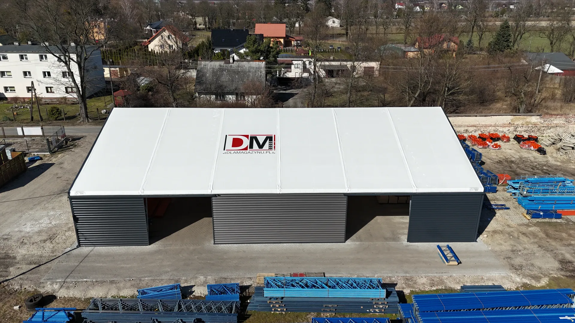 Widok z lotu ptaka na białą, przemysłową konstrukcję namiotową z logo DTM, położoną na utwardzonej działce pośród otaczających ją gruzów.