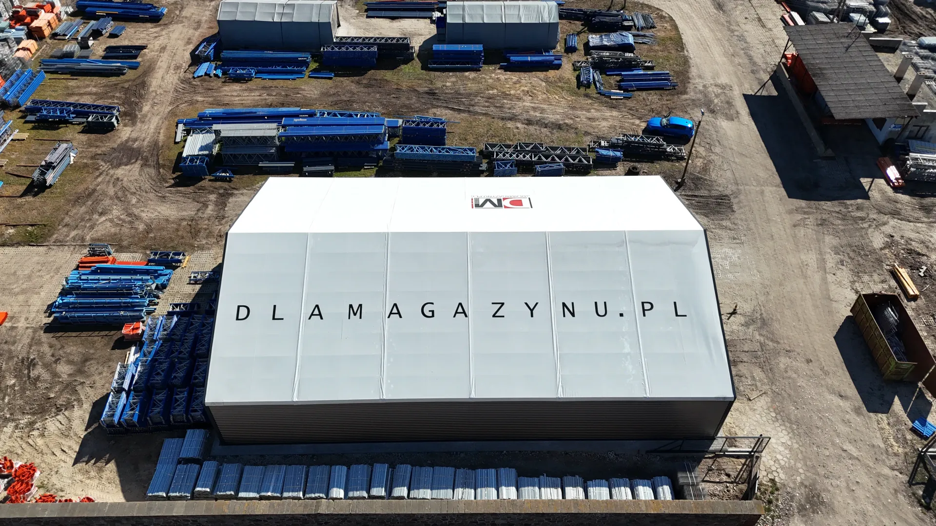 Widok z lotu ptaka na duży, biały namiot magazynowy z napisem „DLAMAGAZYNU.PL” nadrukowanym na dachu, otoczony sprzętem przemysłowym.