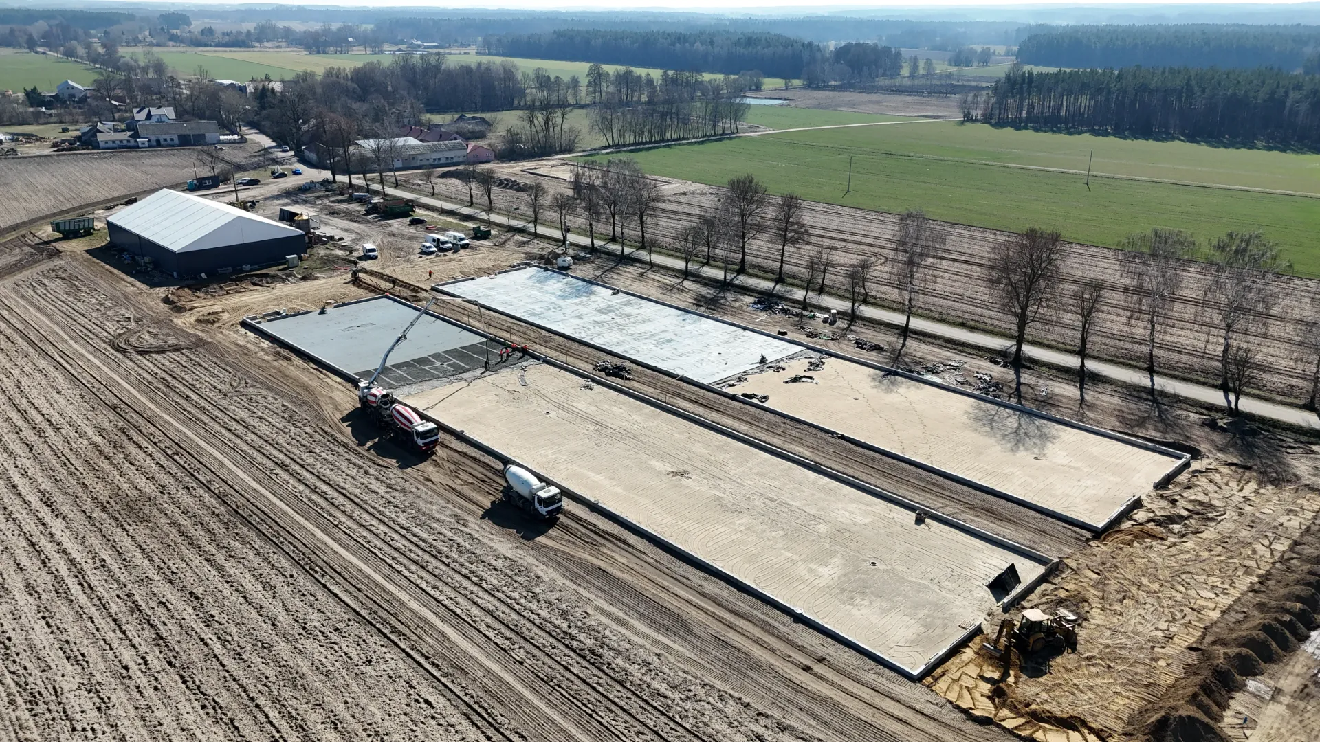 Widok z lotu ptaka na plac budowy, na którym wylewane są betonowe płyty fundamentowe długich budynków na polu wiejskim.