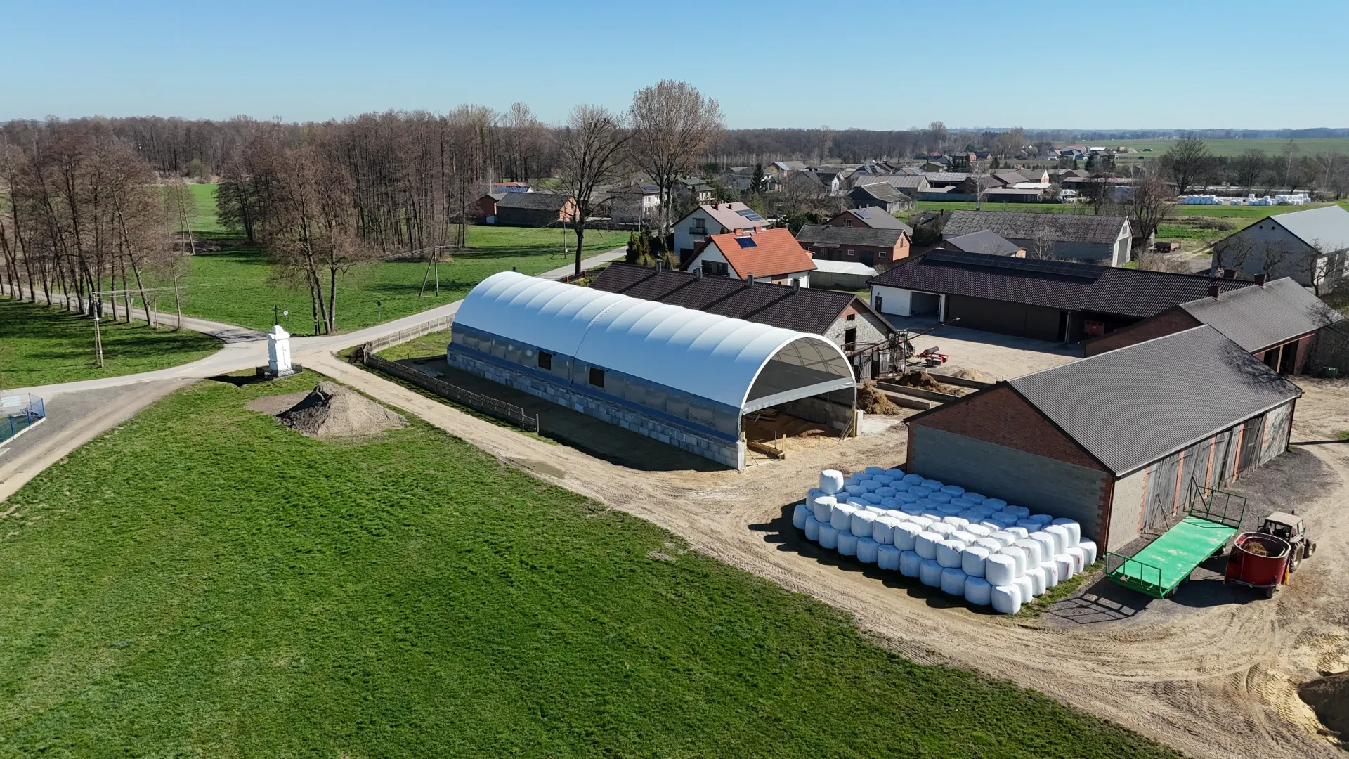 Widok z lotu ptaka na farmę z białym dachem łukowym, brązową stodołą i dużym stosem owiniętych bel siana.