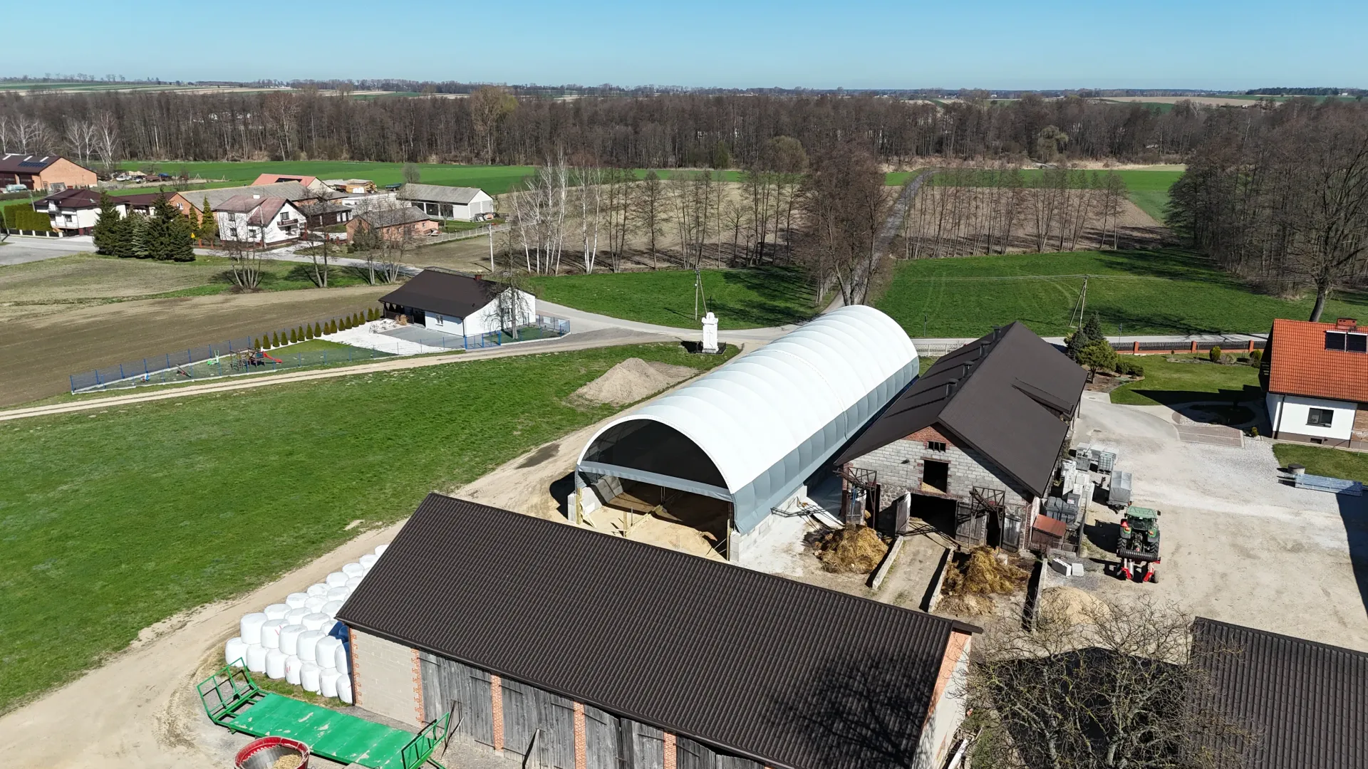 Widok z lotu ptaka na farmę, na którą składa się duża łukowata konstrukcja magazynowa, kilka budynków z ciemnymi dachami i zielone pola.