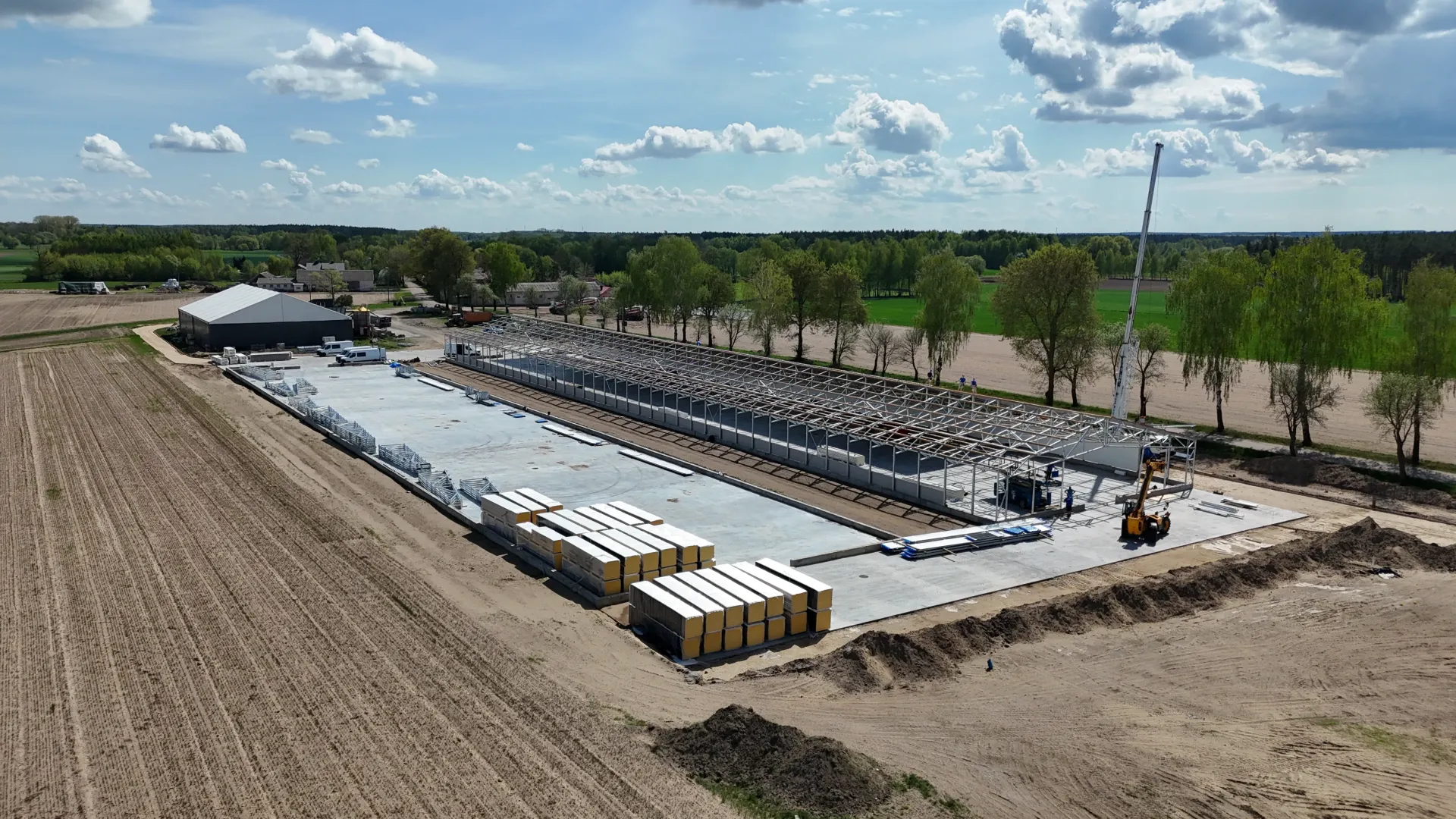 Widok z lotu ptaka na plac budowy z długim betonowym fundamentem, ułożonymi w stos materiałami i dźwigiem na polu wiejskim.