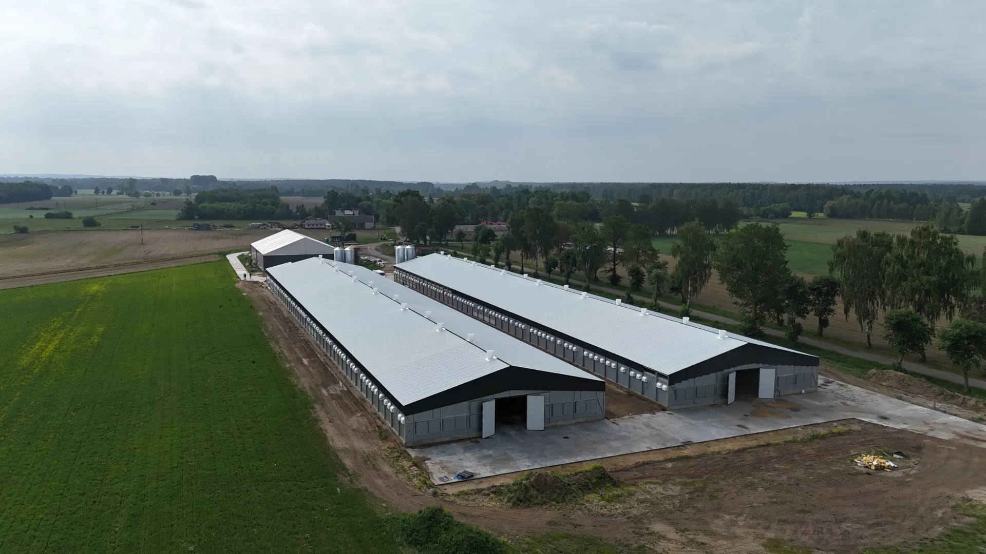 Widok z lotu ptaka na dwa długie, kryte metalowymi dachami budynki magazynowe, służące celom rolniczym, położone na wiejskim polu.