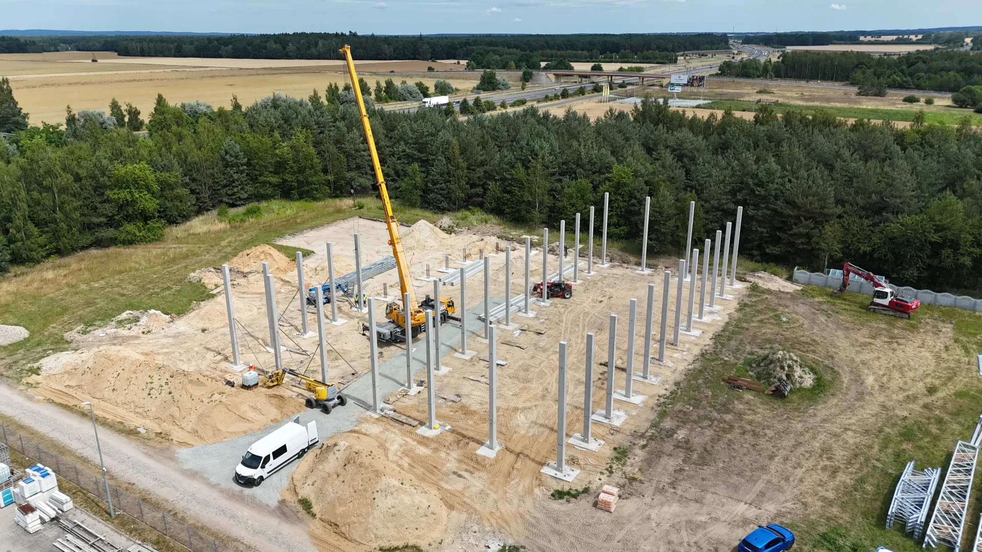 Widok z lotu ptaka na plac budowy, na którym żółty dźwig wznosi betonowe kolumny na piaszczystej działce w pobliżu lasu.