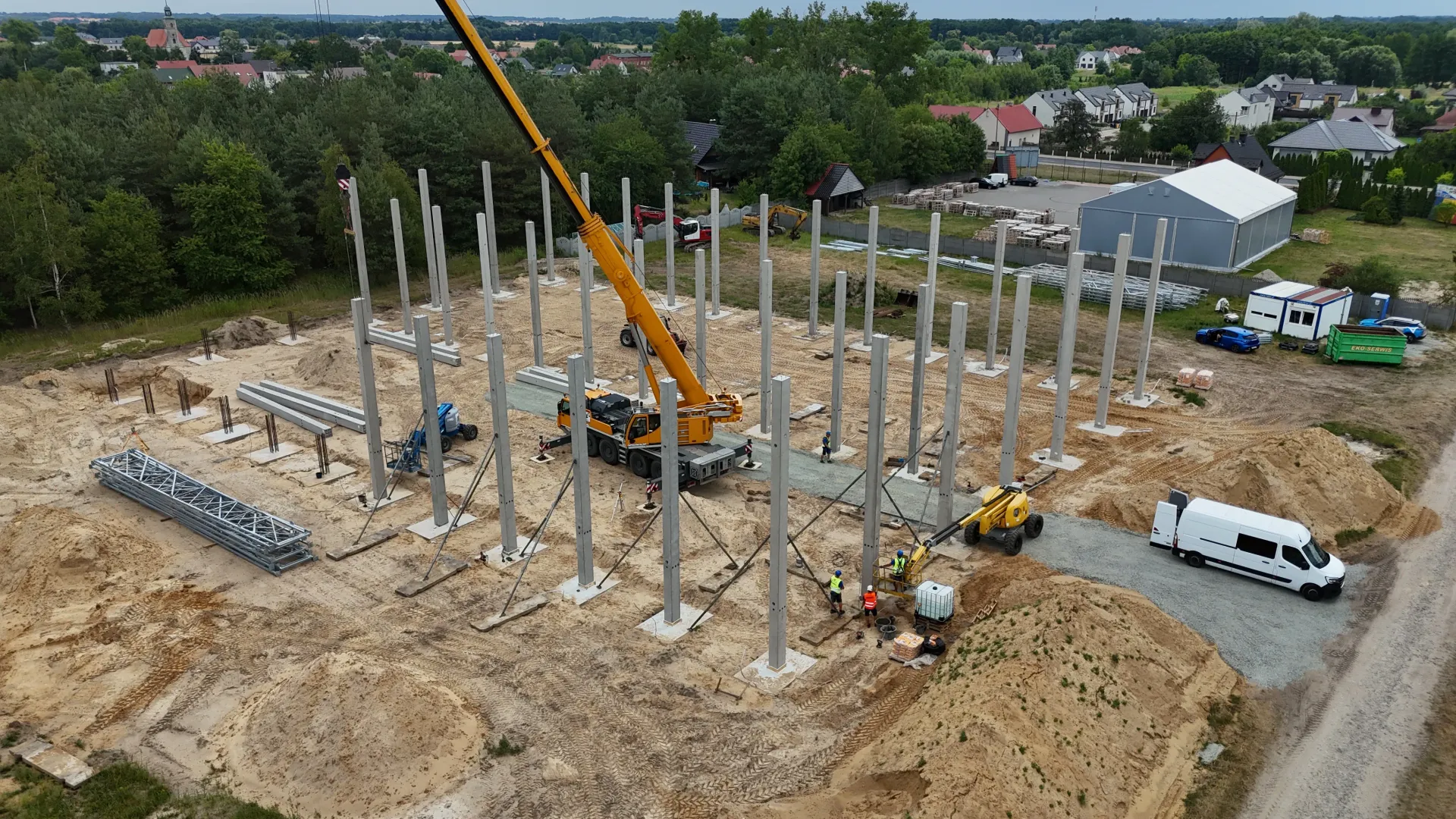 Widok z lotu ptaka na plac budowy, na którym żółty dźwig montuje pionowe betonowe słupy na piaszczystym terenie.