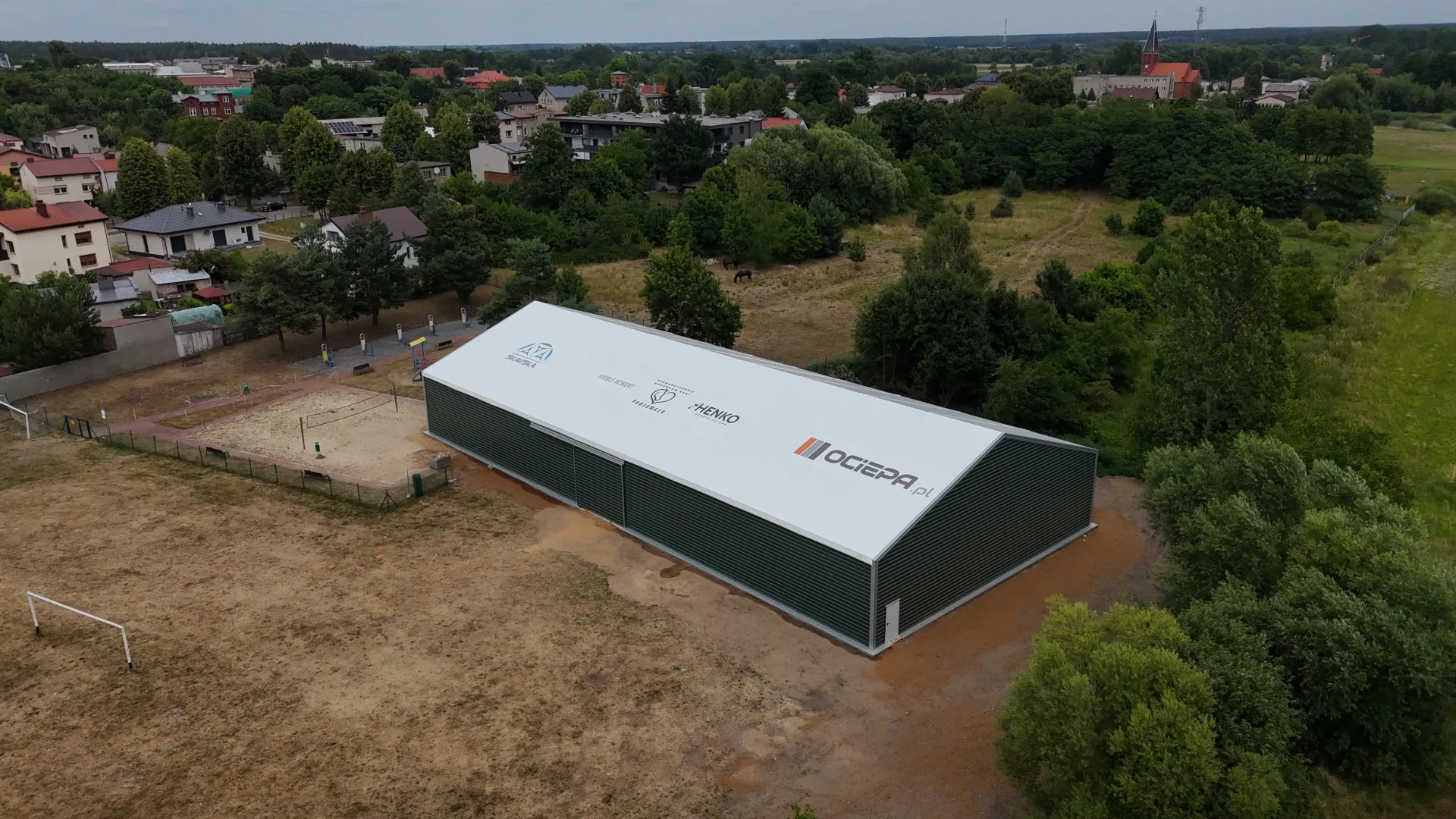 Widok z lotu ptaka na długą, szarą konstrukcję przemysłową, usytuowaną na działce w pobliżu osiedla i drzew.
