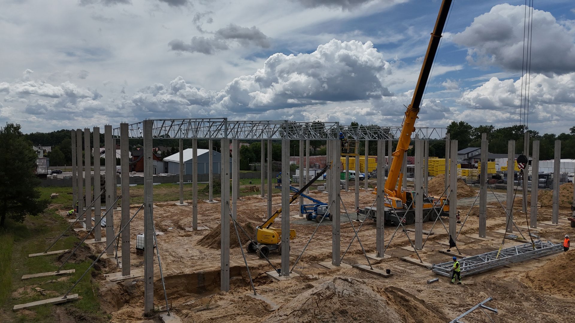 Widok z góry na plac budowy z betonowymi filarami, metalowymi kratownicami dachowymi i żółtym dźwigiem na tle błękitnego nieba.