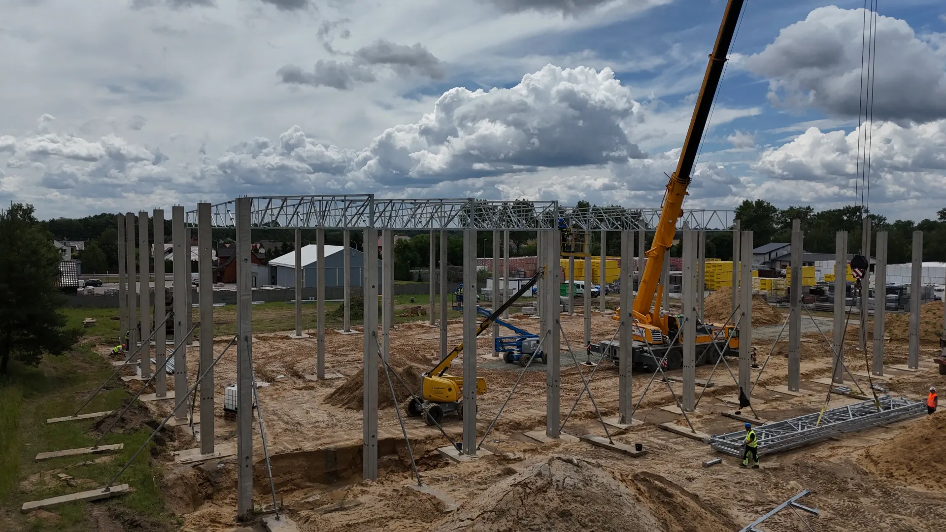 Plac budowy, na którym widać betonowe filary konstrukcji budynku montowane za pomocą żółtego dźwigu na tle zachmurzonego nieba.
