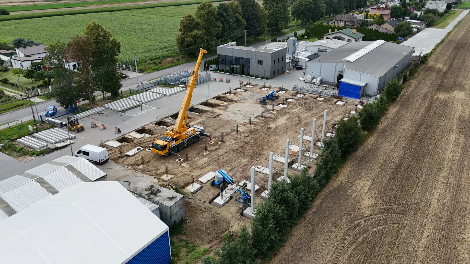 Widok z lotu ptaka na trwający plac budowy, na którym widać żółty dźwig mobilny i pionowe betonowe słupy, na których trwają prace.