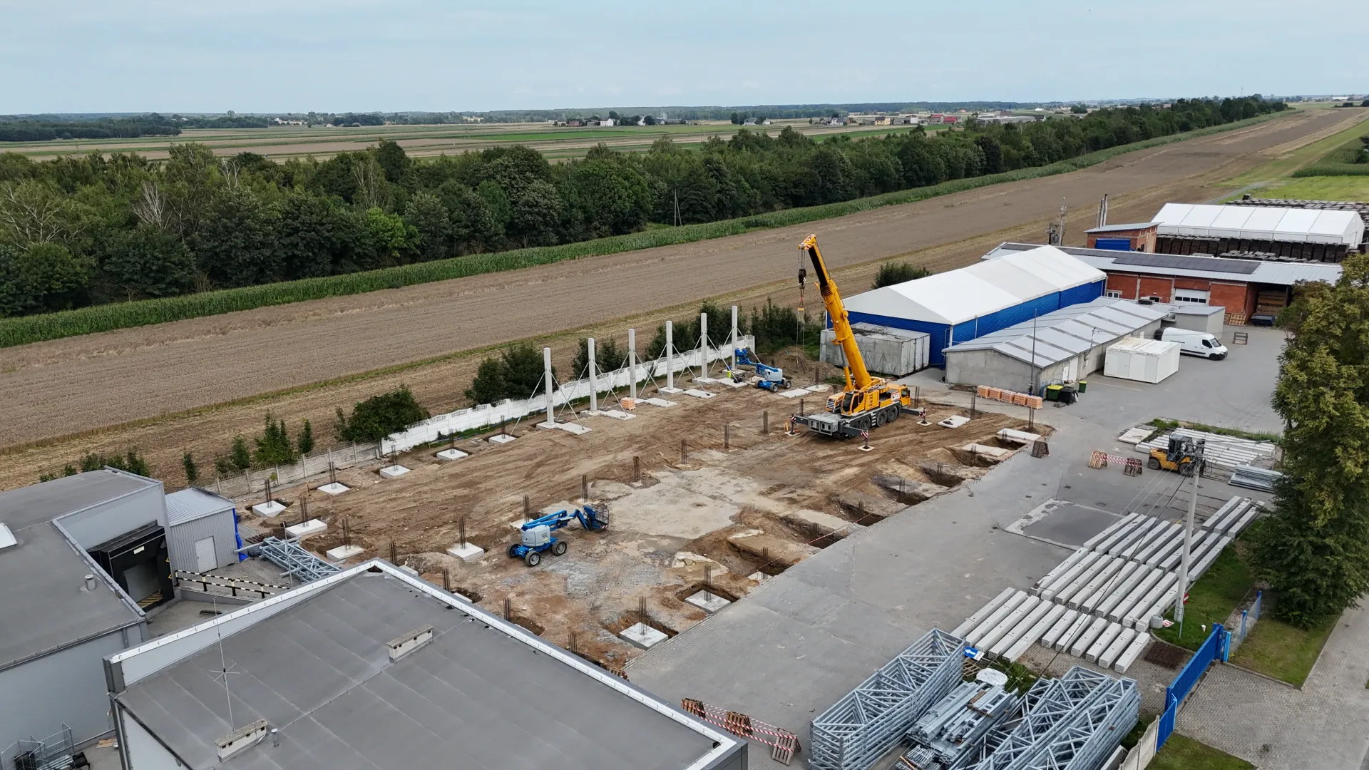 Widok z lotu ptaka na plac budowy z pionowymi betonowymi filarami, żółtym dźwigiem i porozrzucanymi materiałami budowlanymi.