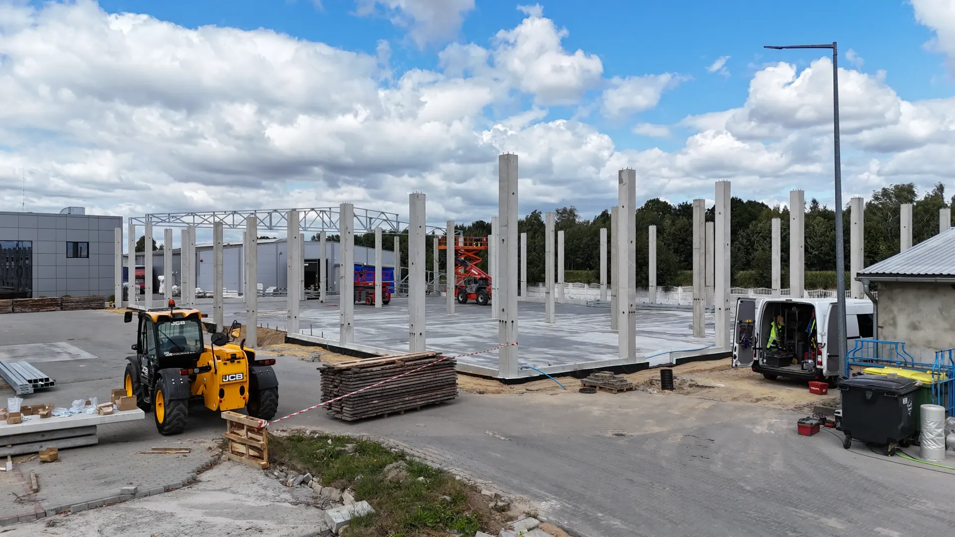 Plac budowy z betonowymi filarami, żółtą ładowarką i białym vanem pod błękitnym niebem z białymi chmurami.