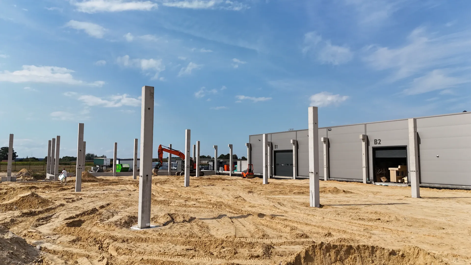 Betonowe filary podporowe stoją na piaszczystym placu budowy przed częściowo ukończonym szarym budynkiem przemysłowym.