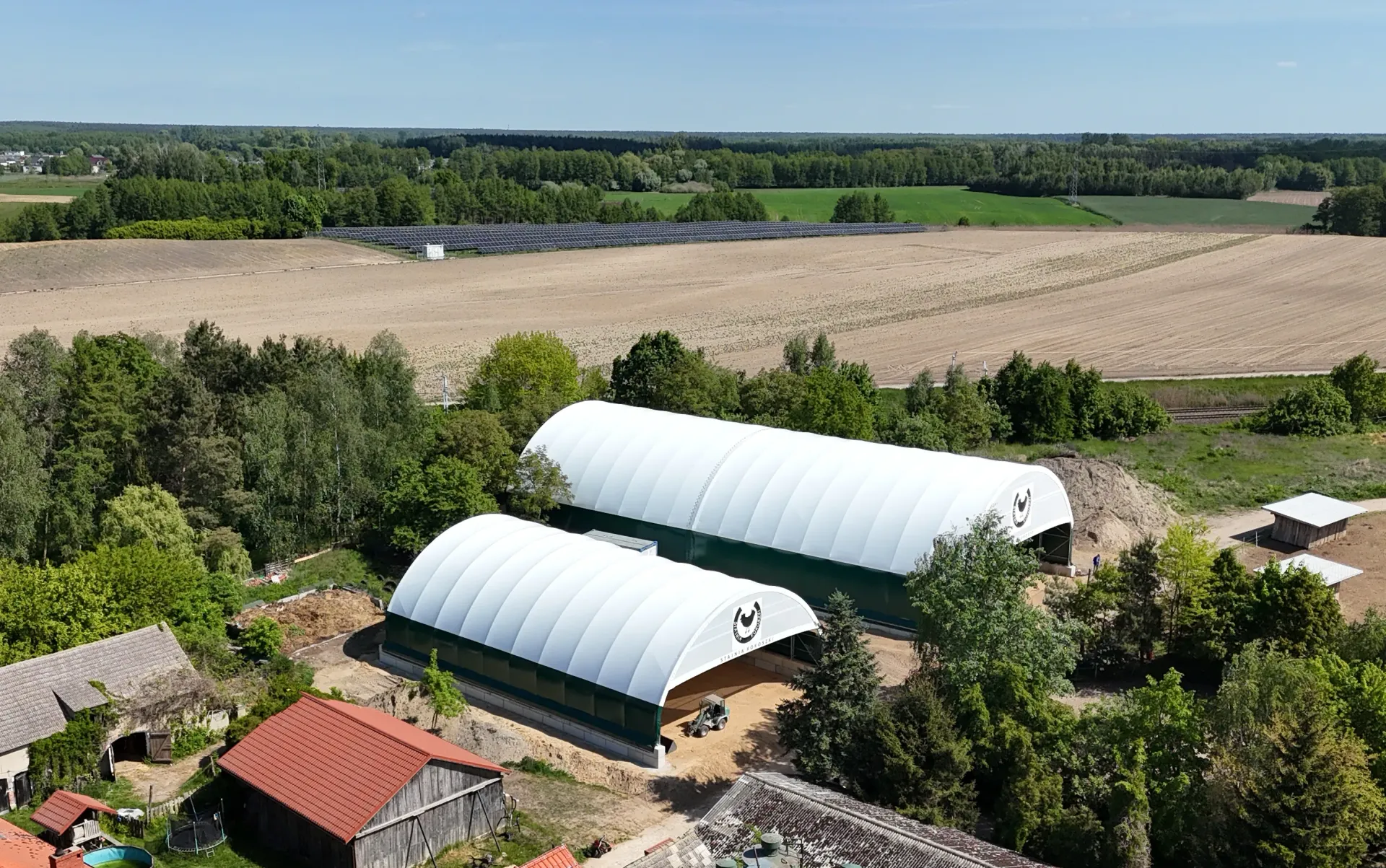 Dwa białe, łukowate budynki z tkaniny znajdują się na tle wiejskiego krajobrazu, wśród drzew, szopy z czerwonym dachem i dużego, otwartego pola.