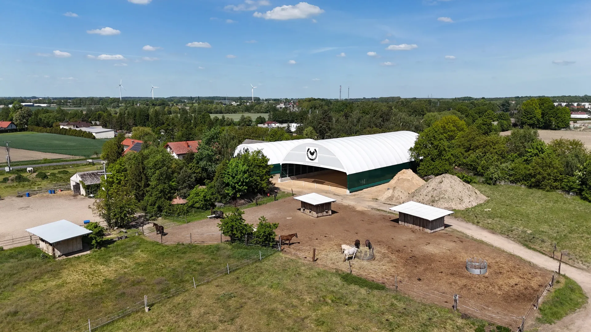 Widok z lotu ptaka na stadninę koni z dużą, białą stodołą, zewnętrznymi padokami i kilkoma końmi pasącymi się na wsi.