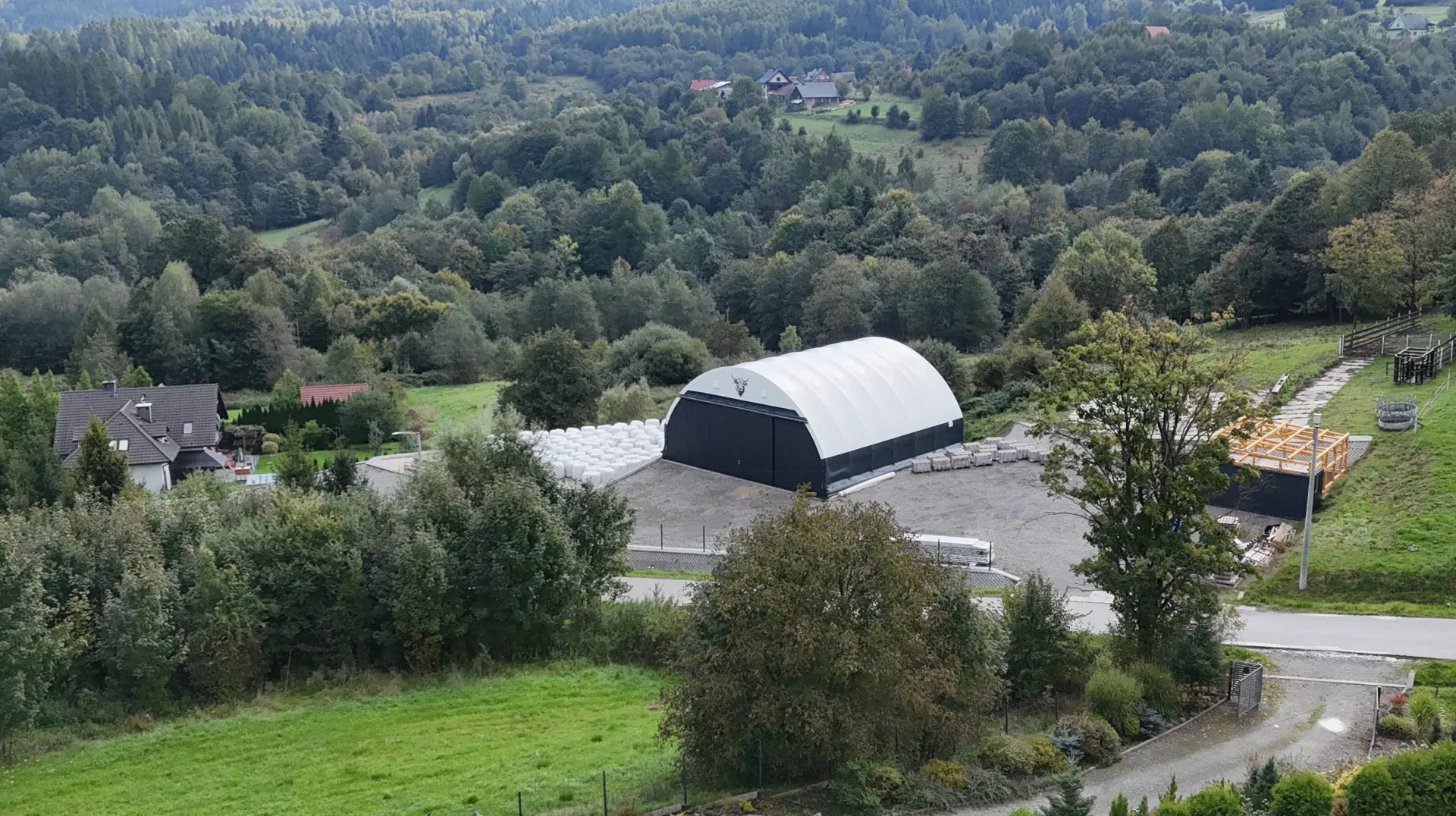 Duża, biała konstrukcja z łukowym dachem i czarnymi ścianami stoi na pokrytym żwirem terenie otoczonym drzewami i zielonymi polami.