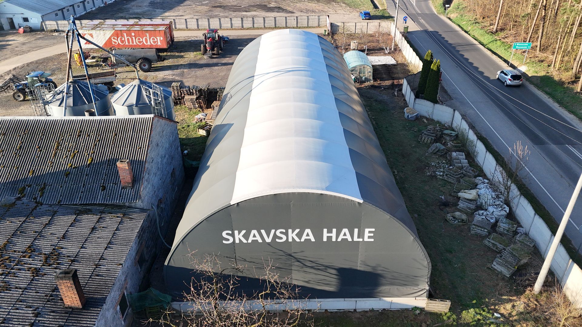 Widok z lotu ptaka na dużą budowlę z dachem łukowym oznaczoną jako „SKAVSKA HALE”, położoną przy drodze i polach uprawnych.