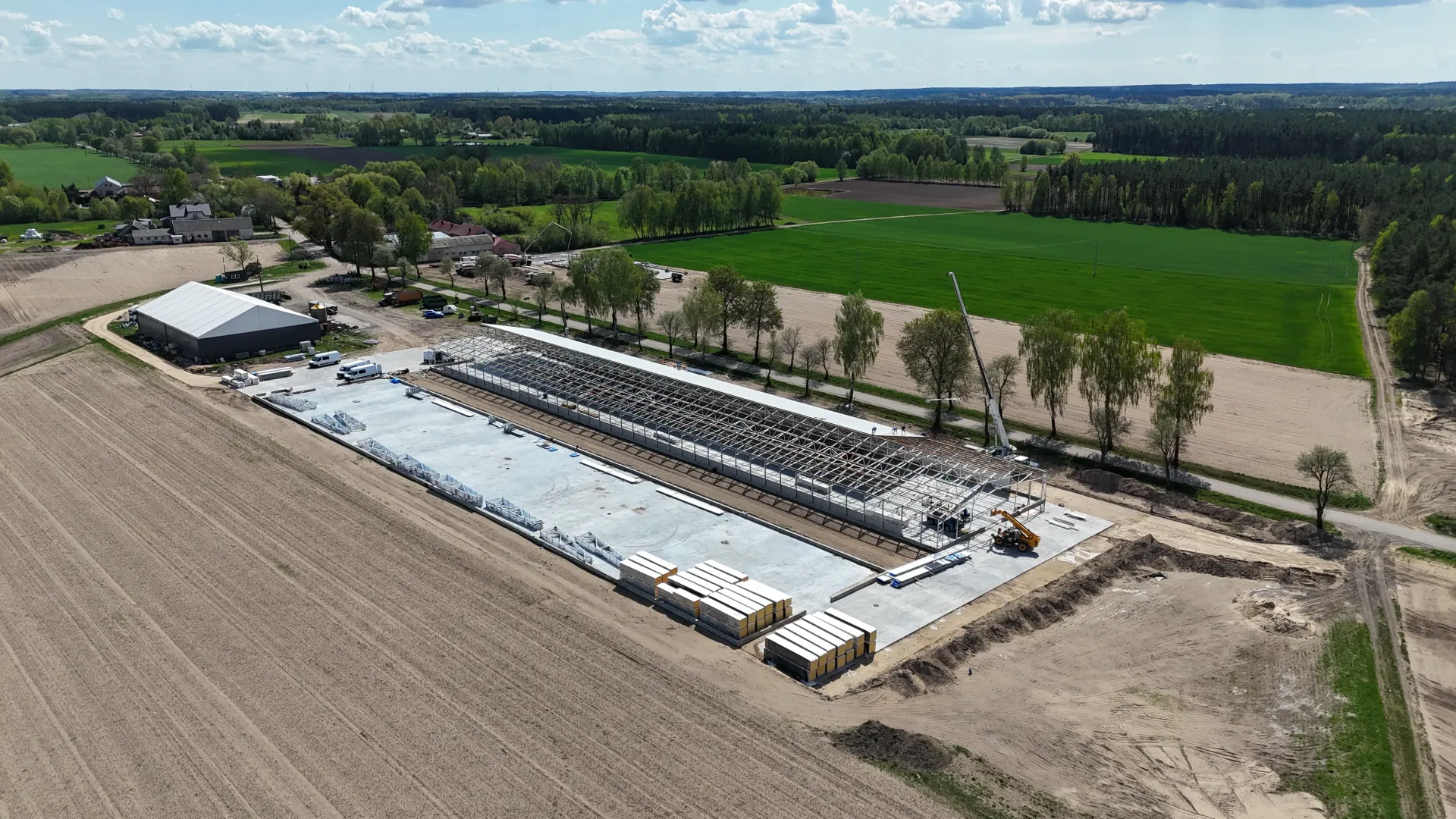 Widok z lotu ptaka na prostokątny plac budowy z betonowym fundamentem, ułożonymi w stos materiałami i otoczeniem pola.