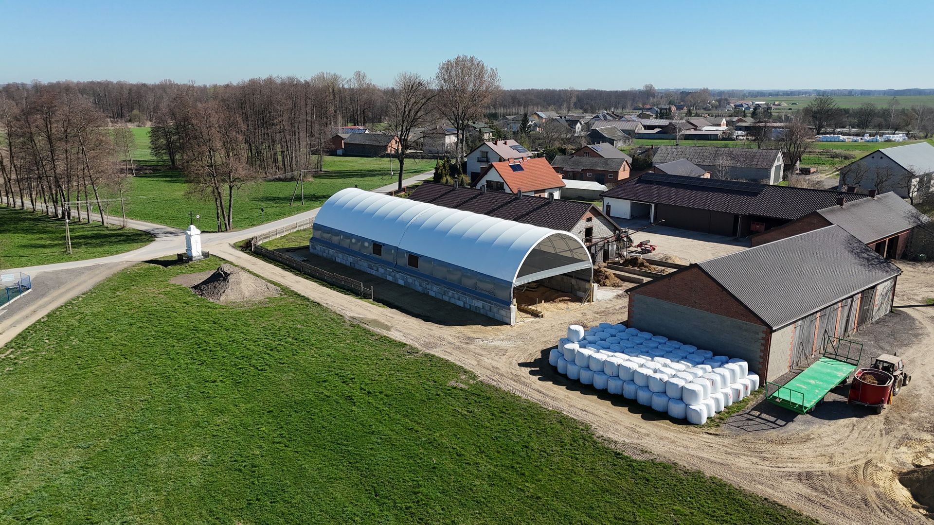 Widok z lotu ptaka na farmę z białym, łukowatym schronieniem, przyległymi kamiennymi budynkami i stosami owiniętych bel siana.