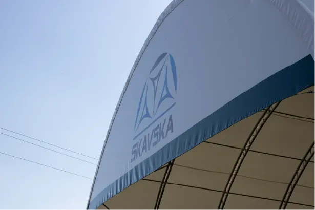 Zbliżenie na zewnętrzną część białego, łukowatego namiotu z niebieskim logo firmy i napisem „SKANSKA” na tle czystego, błękitnego nieba.