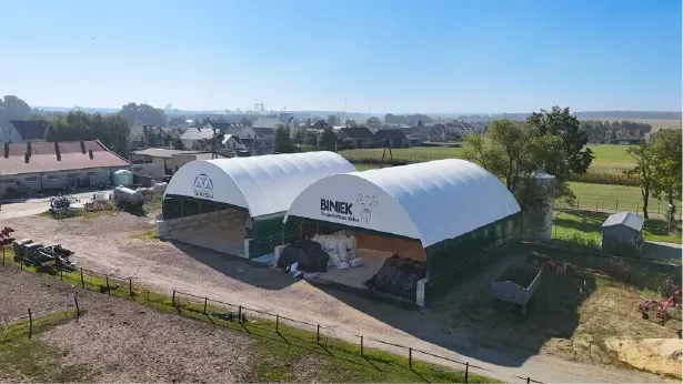 Dwie duże, białe, łukowate konstrukcje z tkaniny oznaczone „B-NEX” stoją na wiejskim terenie pełnym stosów materiałów i sprzętu.