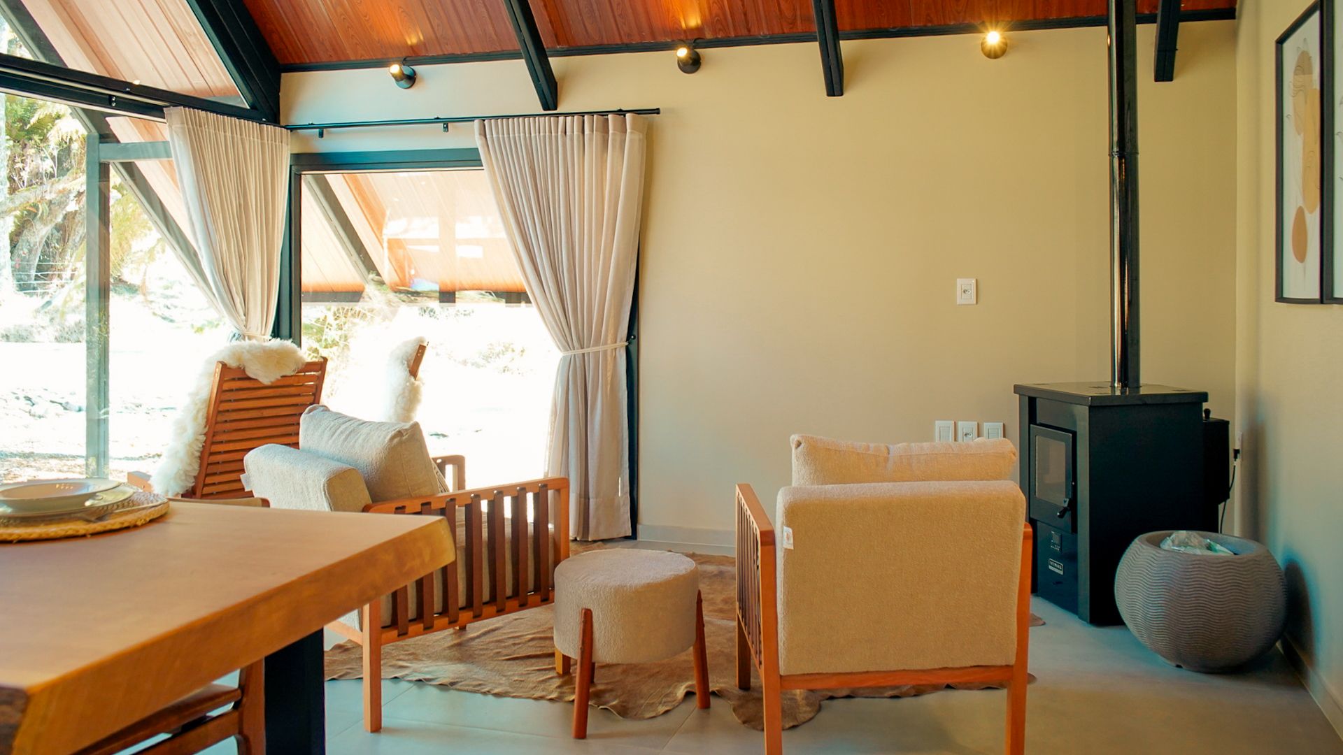 Interior aconchegante da cabana com móveis de madeira, lareira e janelas amplas com vista para a vegetação. Paleta de cores neutras.