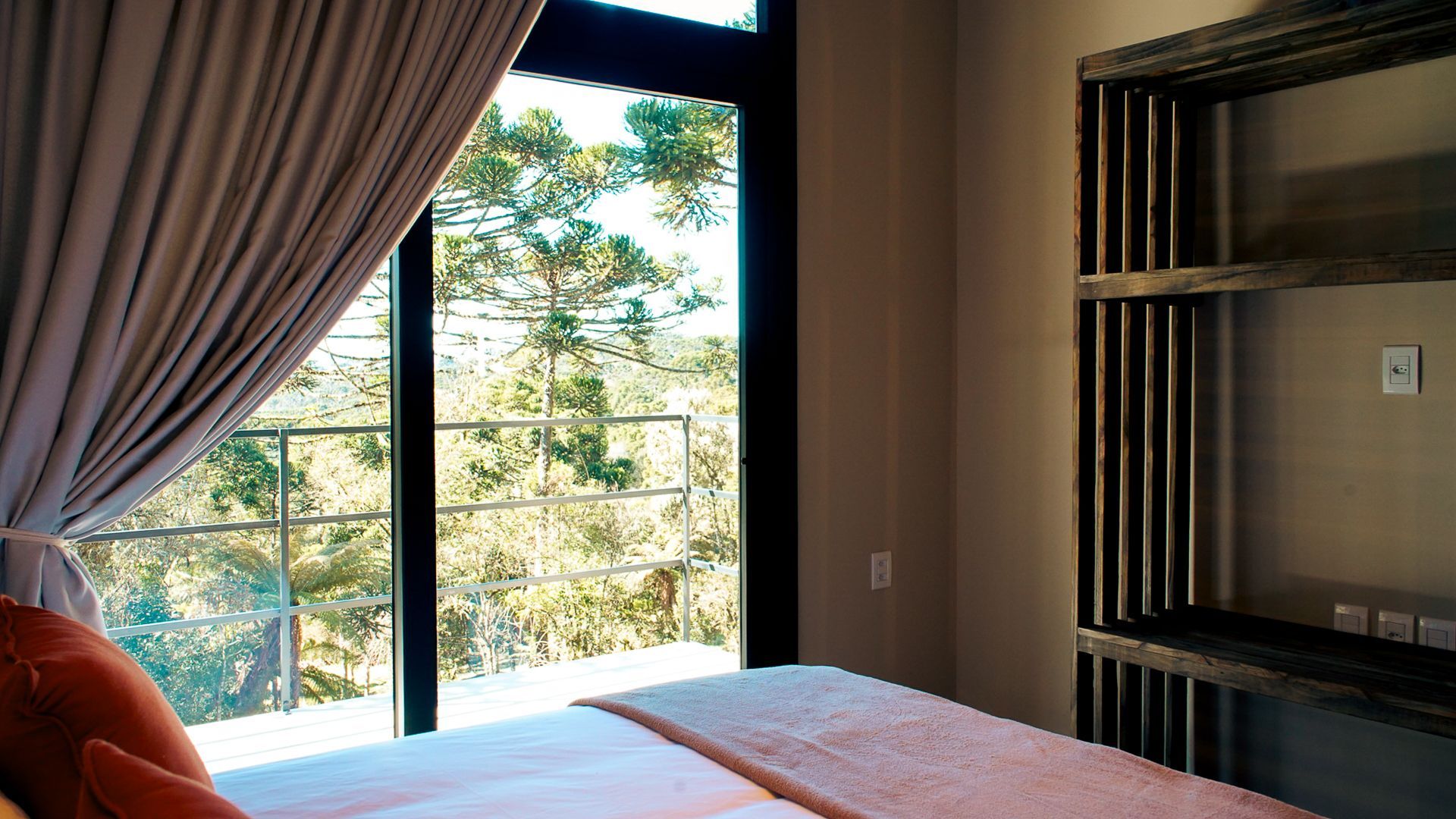 Interior do quarto com uma grande janela com vista para o verde. Tons neutros, cama visível, estante aberta à direita.