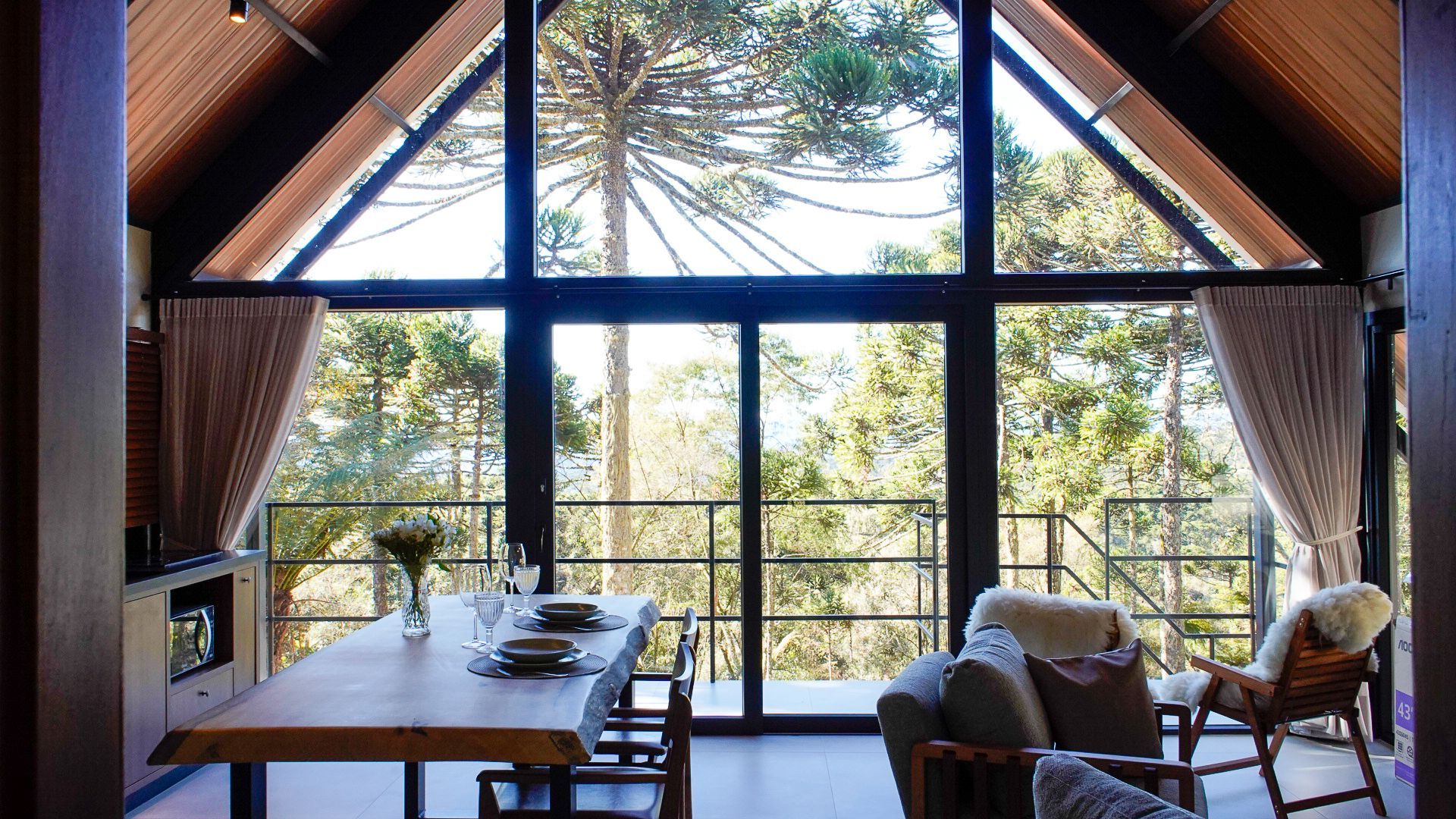 Interior de uma cabana com uma grande janela com vista para uma floresta. Mesa de jantar e assentos visíveis.