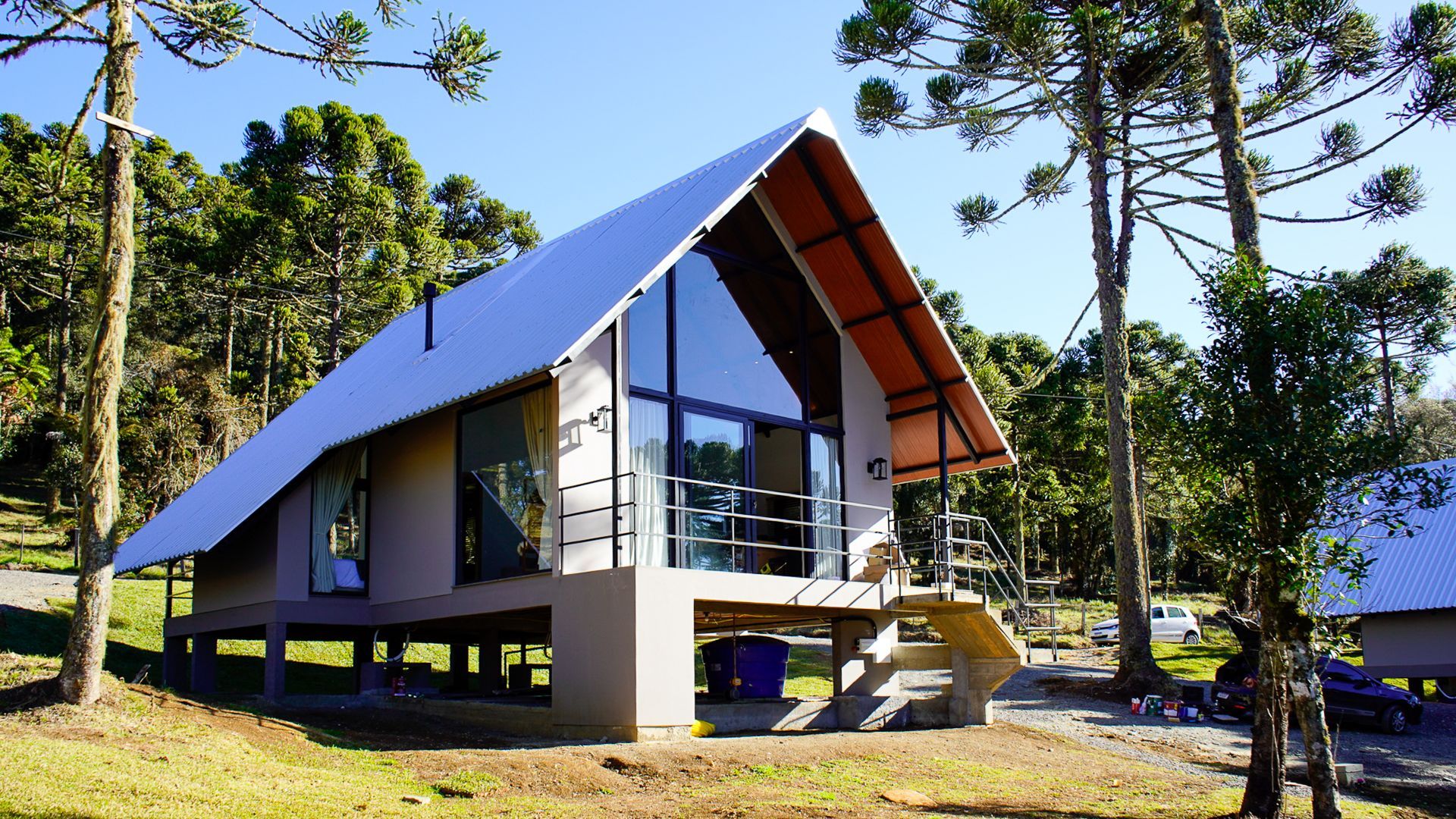 Uma cabana moderna com teto inclinado e grandes janelas sobre palafitas em uma área arborizada.