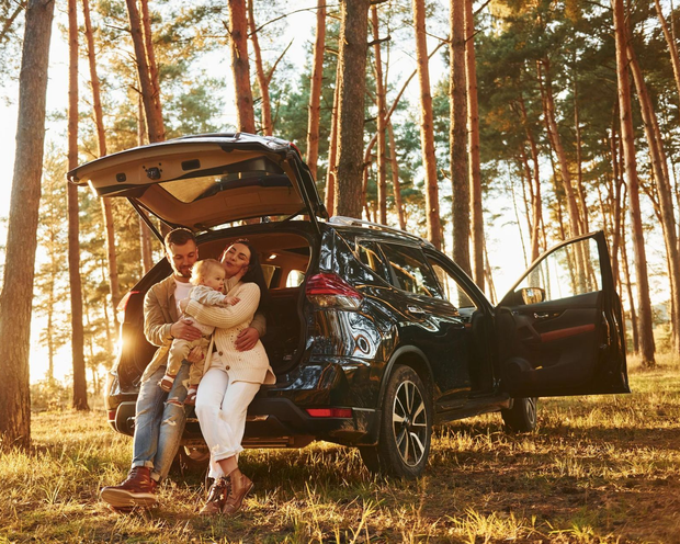Couple sitting on an SUV tailgate in a sunlit forest, with the trunk and door open.