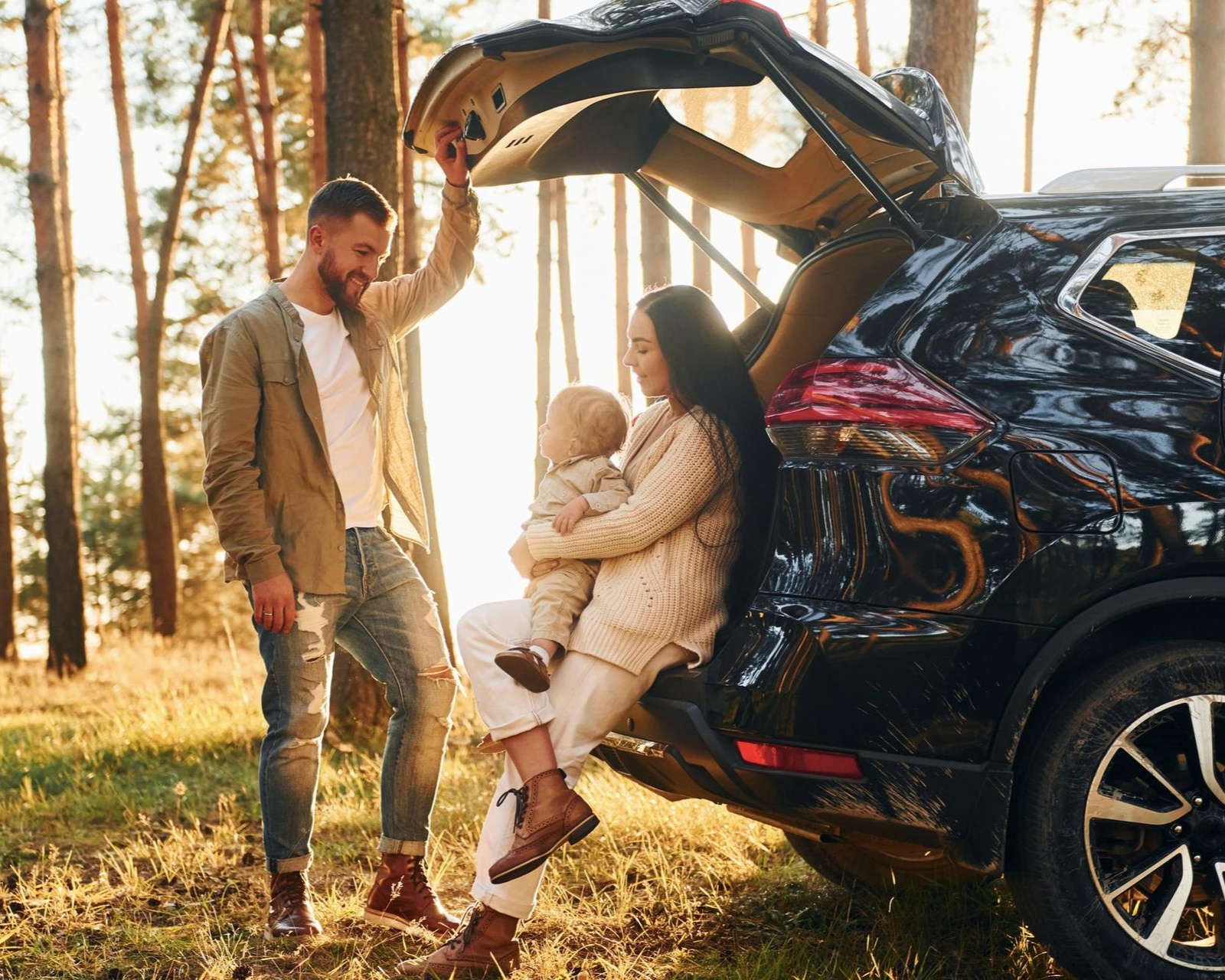 Couple by open SUV in a sunlit forest, one sitting in the trunk while the other stands nearby