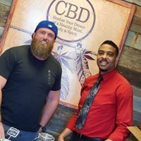Two men smiling indoors beside a CBD sign, one in a black shirt and blue cap, the other in a red shirt and tie
