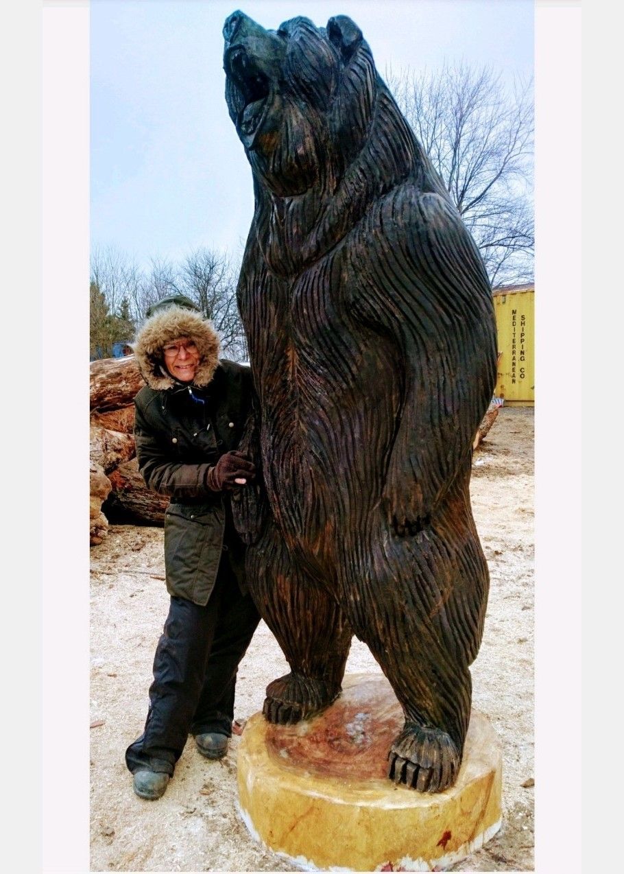 Person standing beside a large wooden bear statue outdoors in winter, on a snow-covered ground.