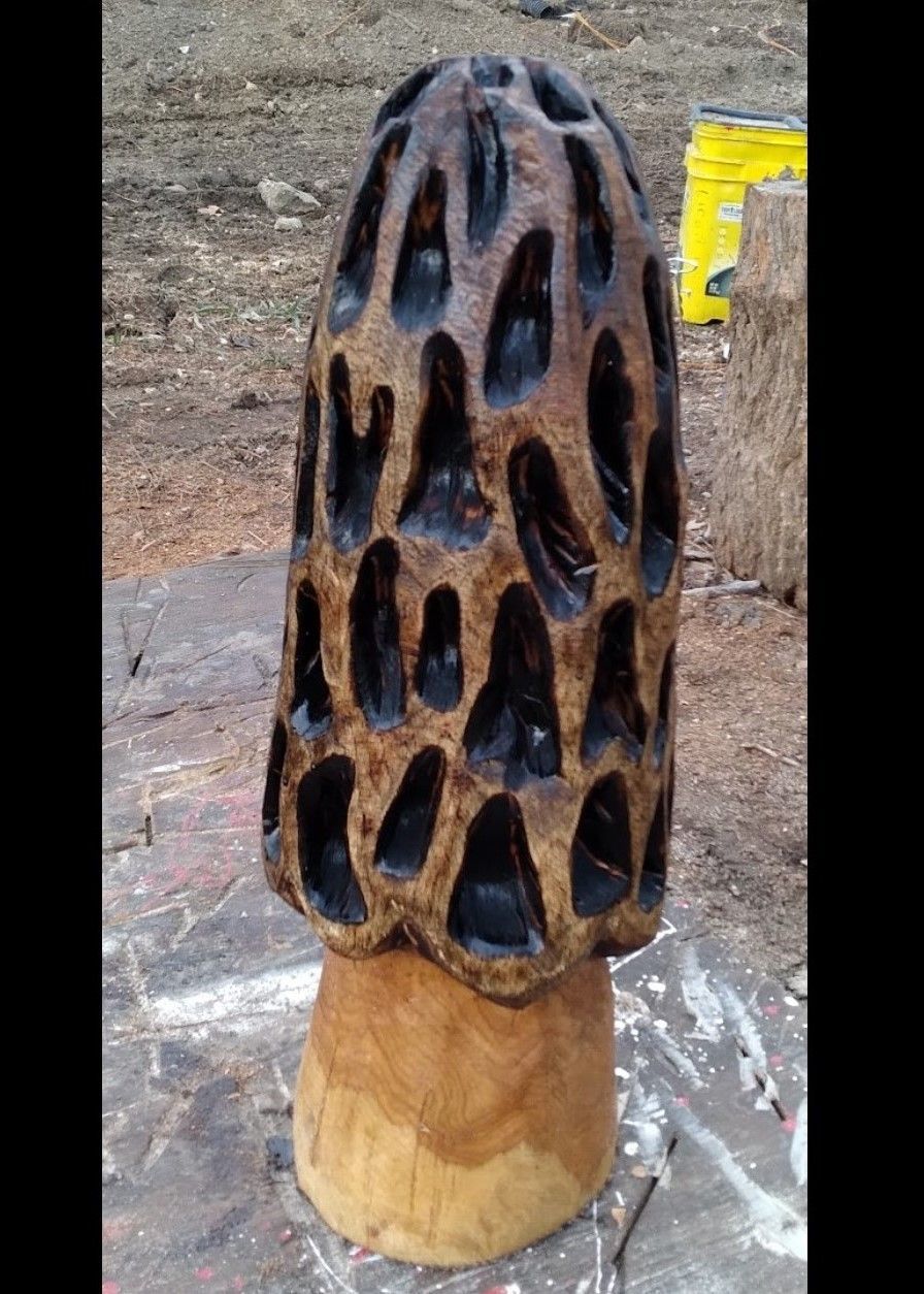Dark carved wooden sculpture resembling a pinecone on a light wood base outdoors