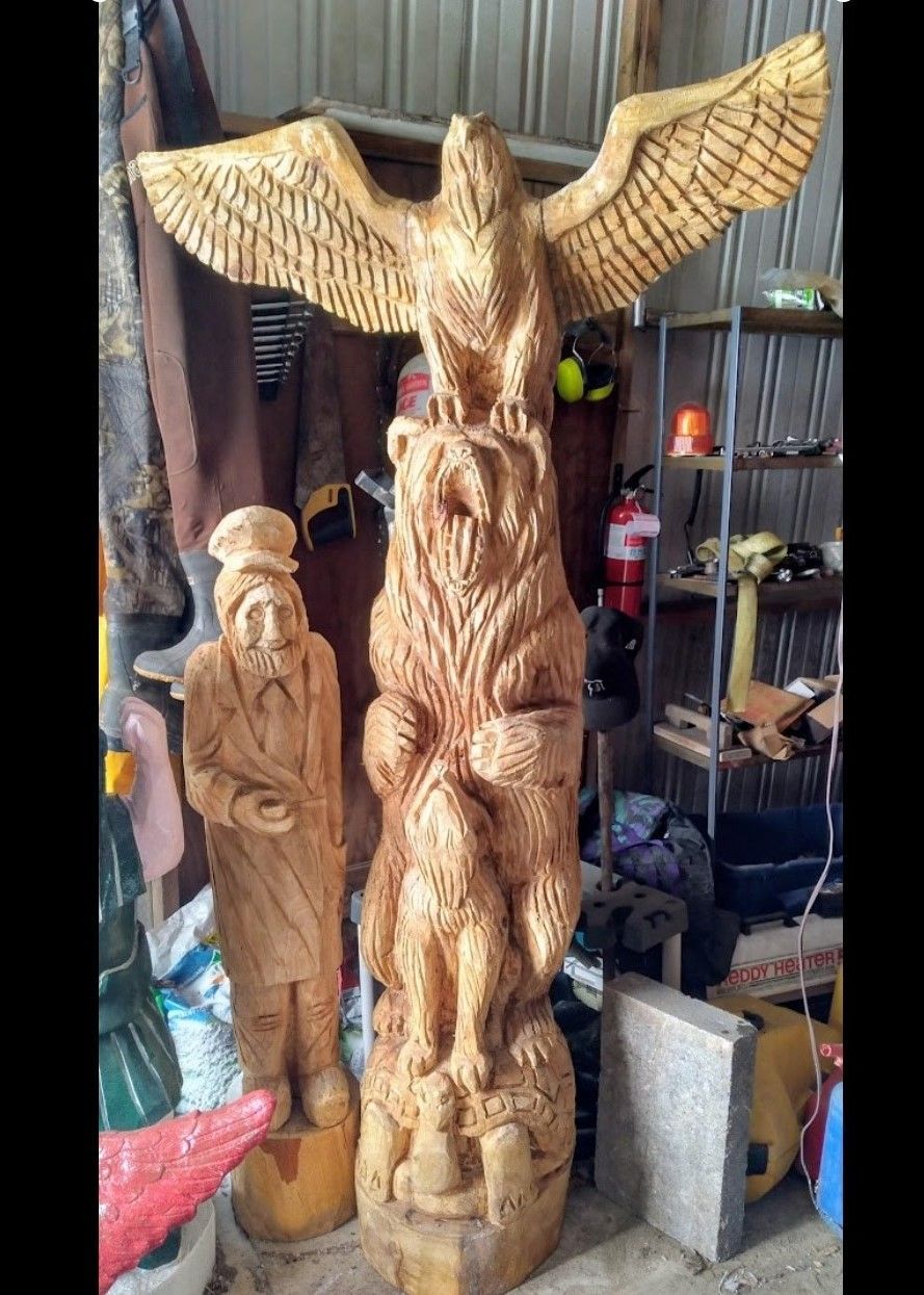 Tall carved wooden angel sculpture with outstretched wings in a workshop, beside another carved figure.