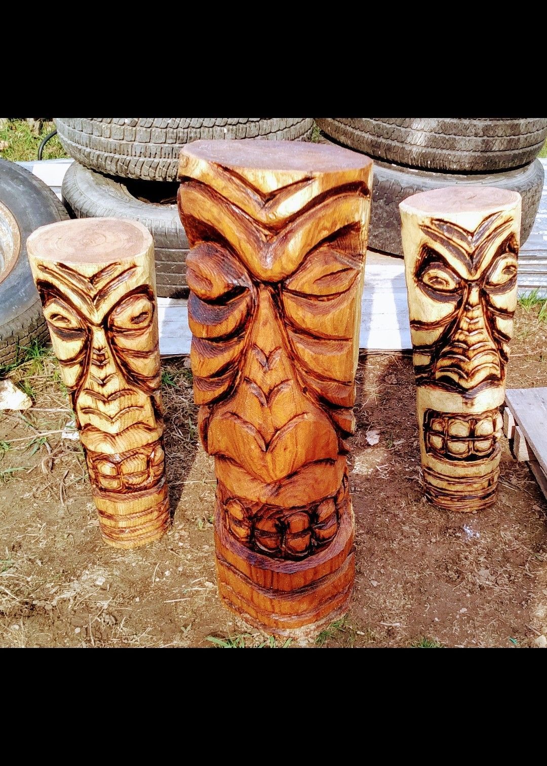 Three carved wooden tiki totem poles in a stone outdoor display, with a large orange-brown center pole.