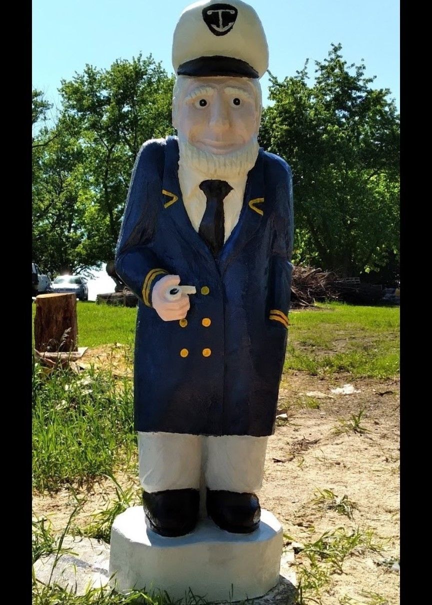 Statue of a sailor in a blue coat and hat standing on a pedestal outdoors in a grassy area.
