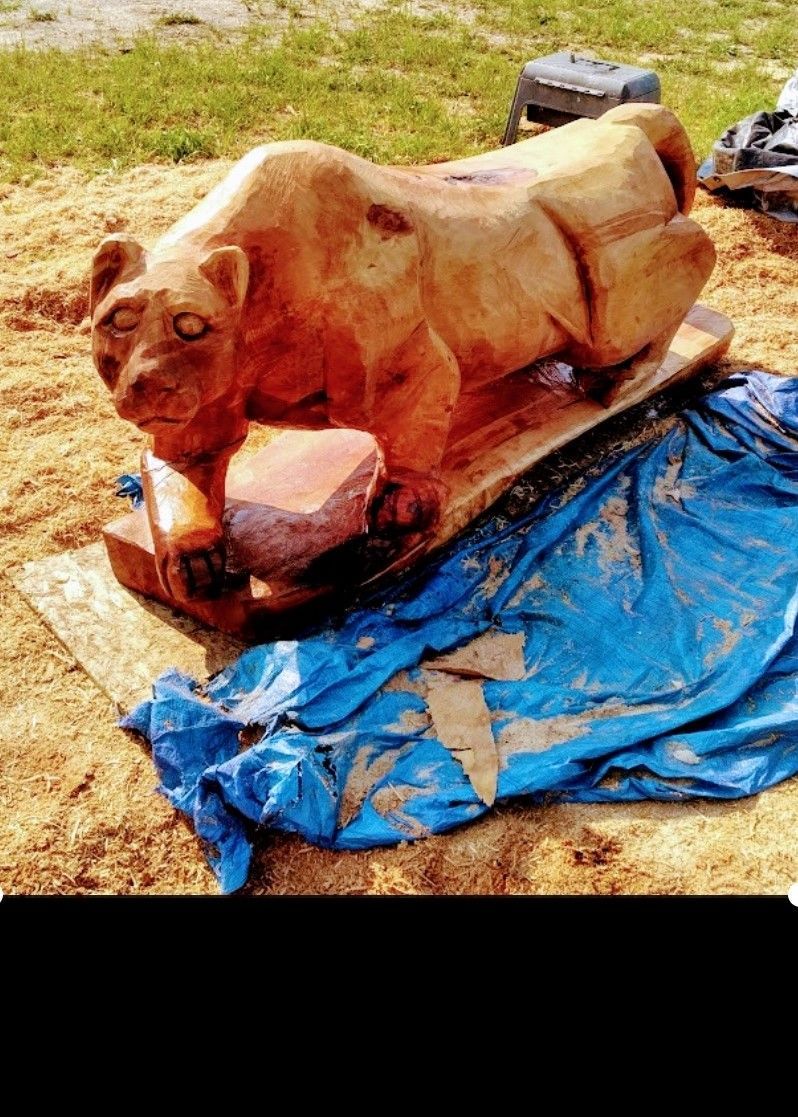 Wooden animal sculpture on a blue tarp outdoors, with reddish-brown paint and grassy background