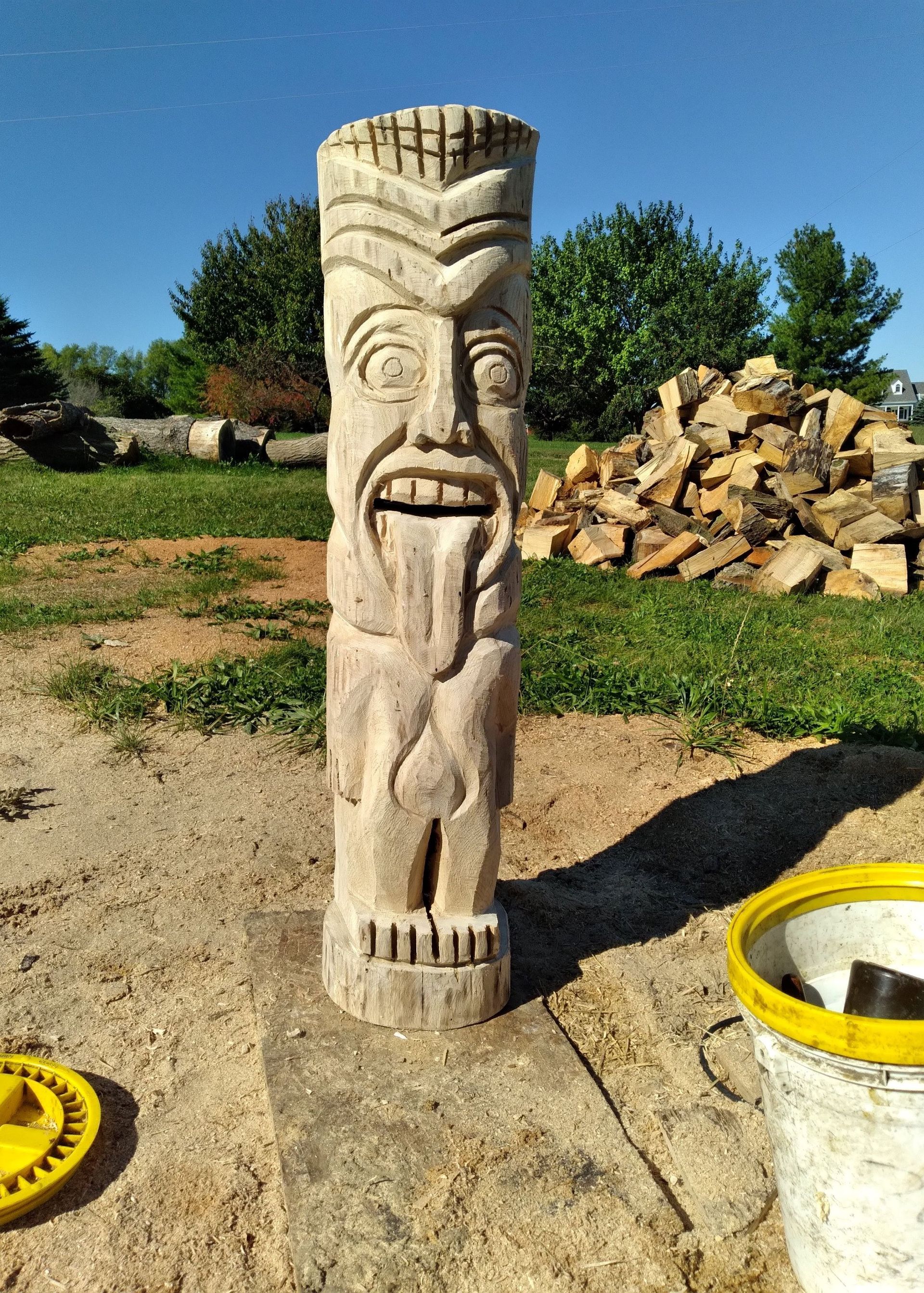 Carved wooden tiki statue outdoors on a sunny gravel path, with stacked stones and greenery behind it