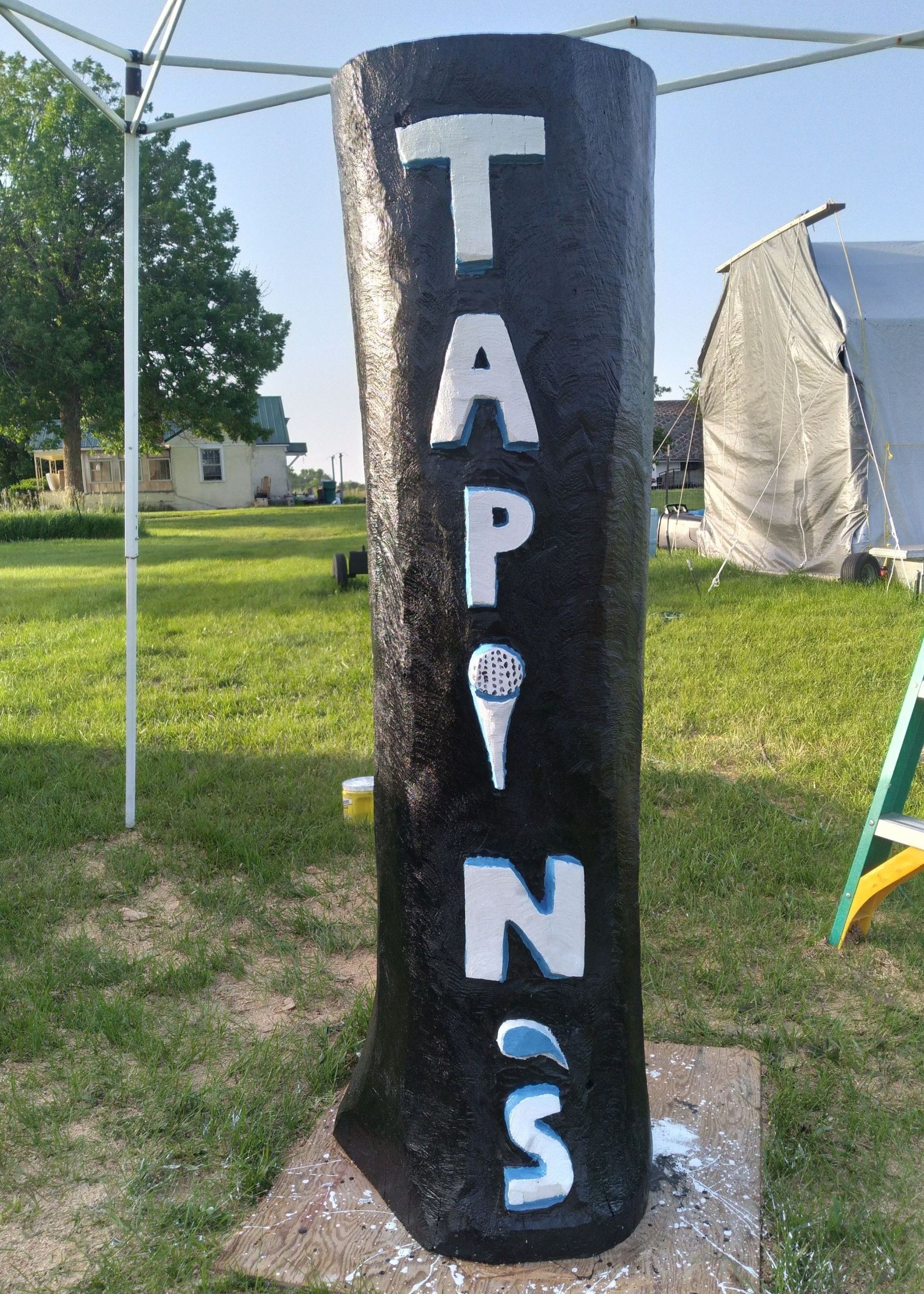 Black painted post with white vertical letters “TAPNS” on grass outdoors.