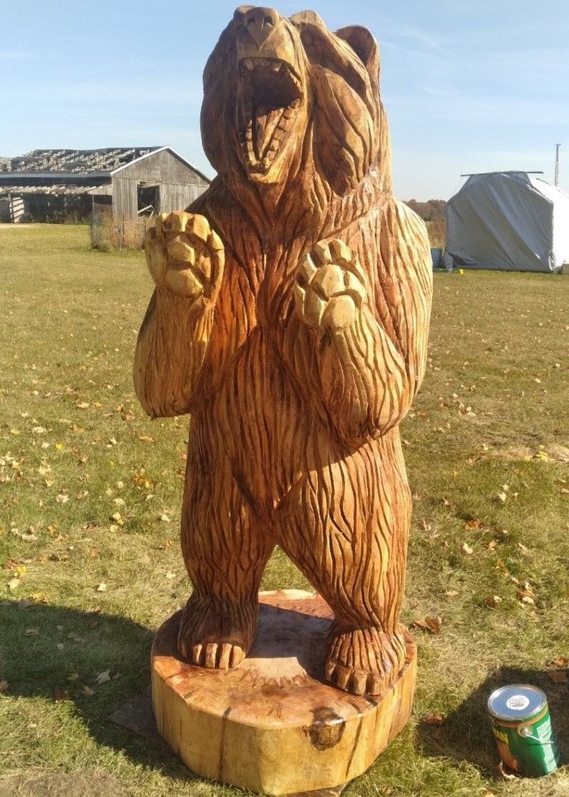 Wooden bear carving standing on a log base in a grassy field, with tents and a small building in the background.
