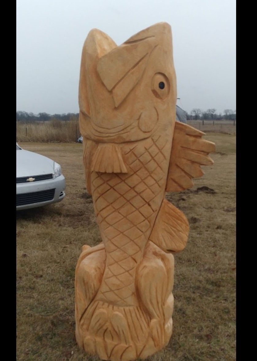 Tall carved wooden fish sculpture standing outdoors in a grassy field near a car.